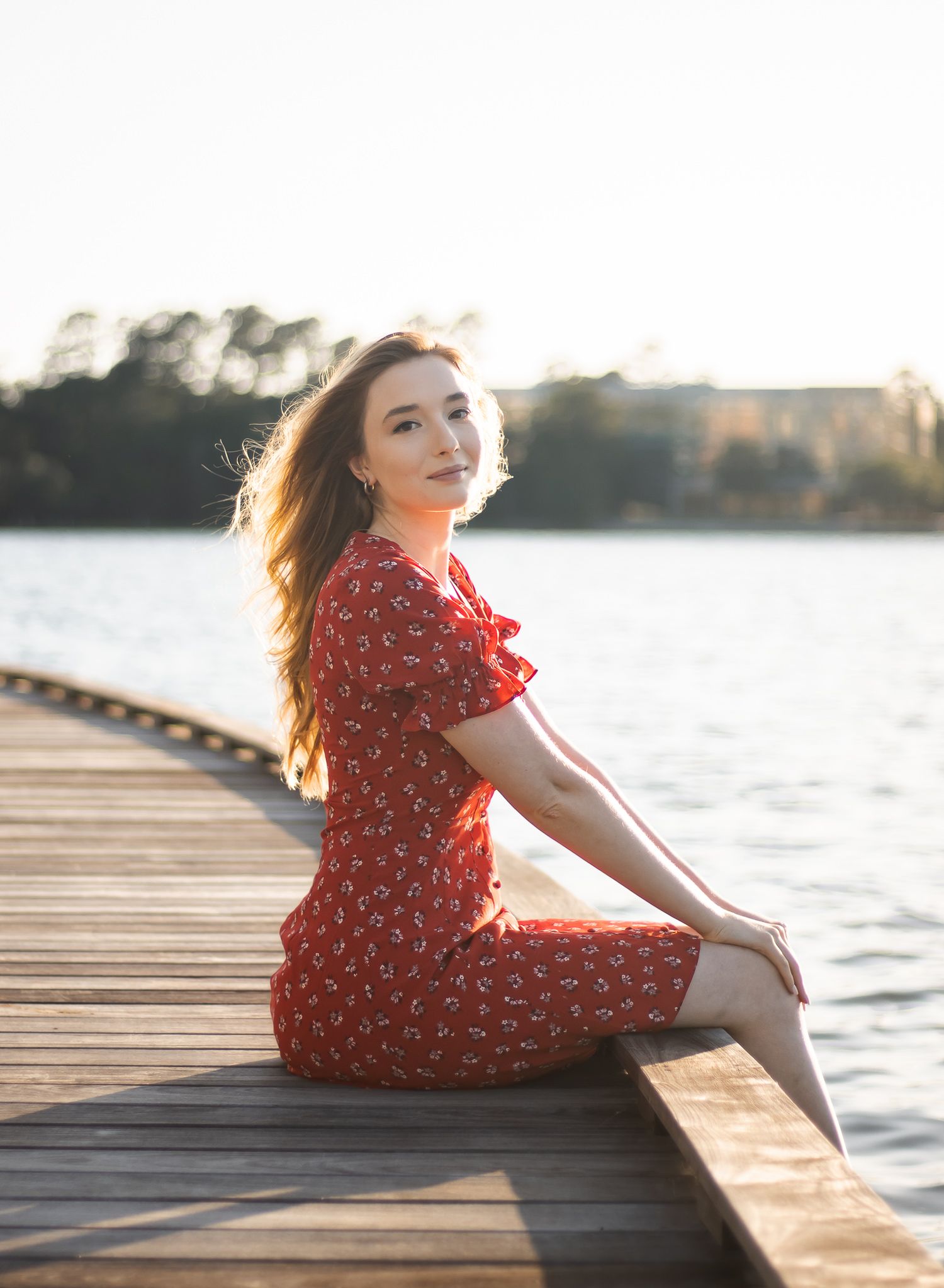 Young woman in red floral dress sits on wooden dock by sunlit lake.