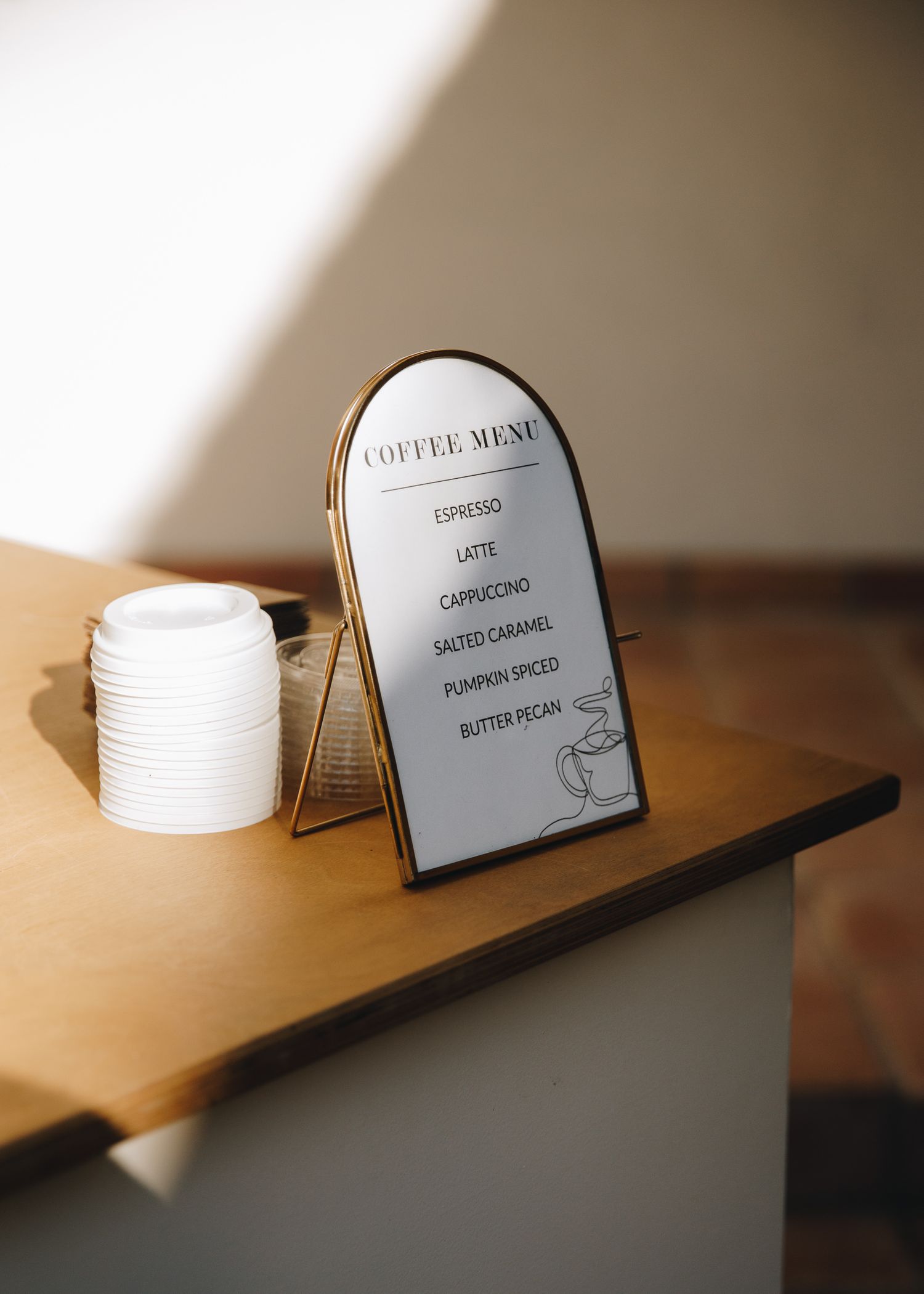Gold-framed arched coffee menu sign displaying espresso, latte, cappuccino, and seasonal drinks on a wooden table.