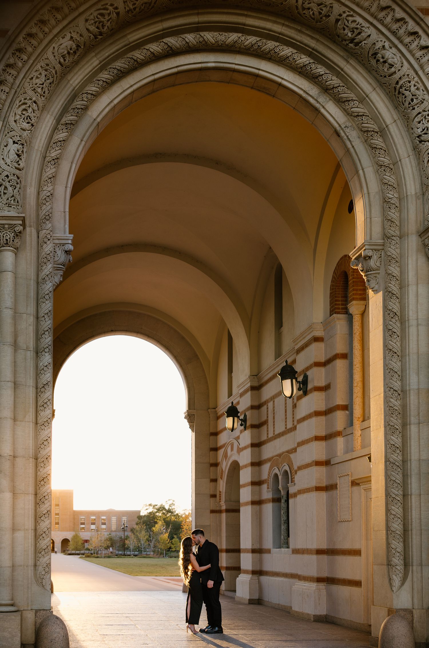 Couple sharing a romantic moment under grand stone arches in a warmly lit historic building corridor.