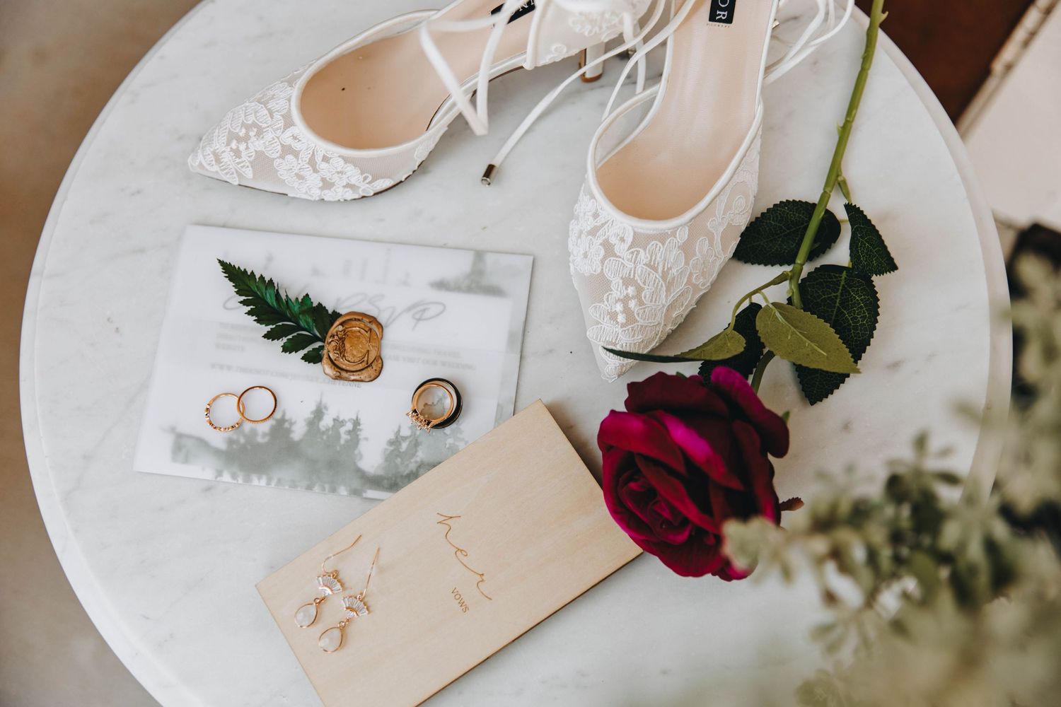 White lace bridal heels, gold rings, wax seal, red rose, and wedding stationery arranged on a marble table.