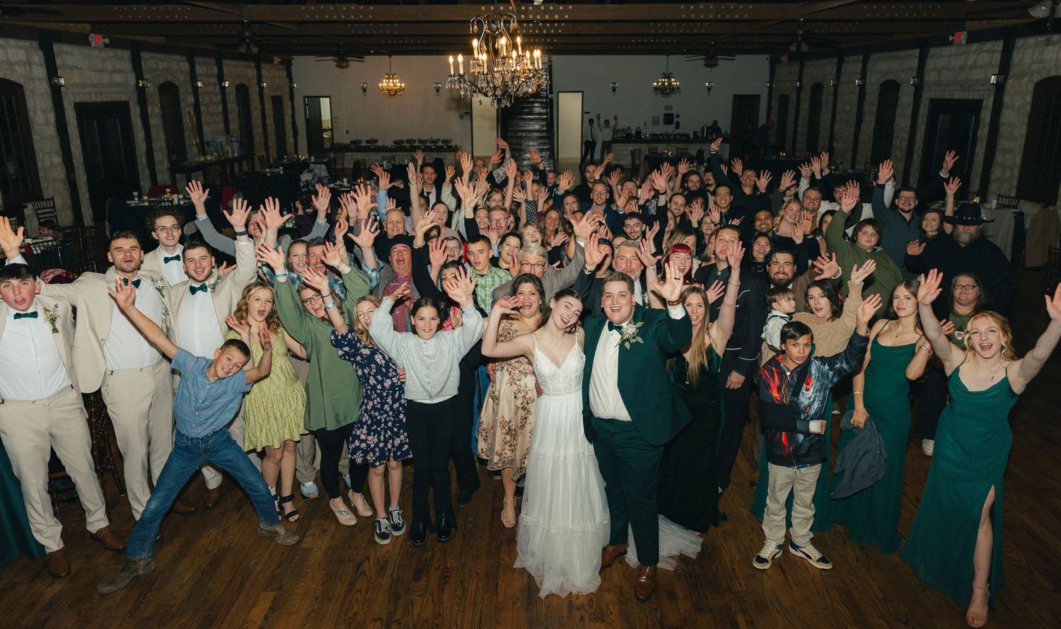 Bride, groom, and wedding guests celebrate with hands raised in grand ballroom group photo at reception.
