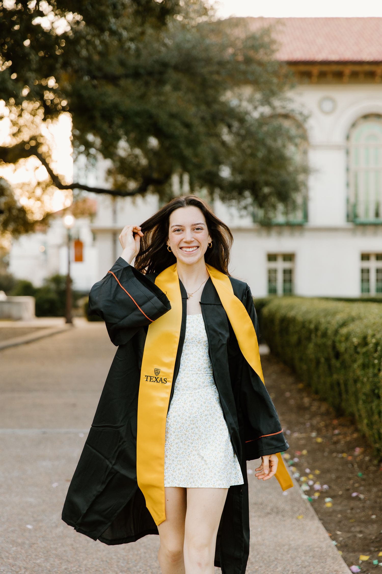 Smiling University of Texas graduate in black gown and gold stole walking on campus at golden hour.