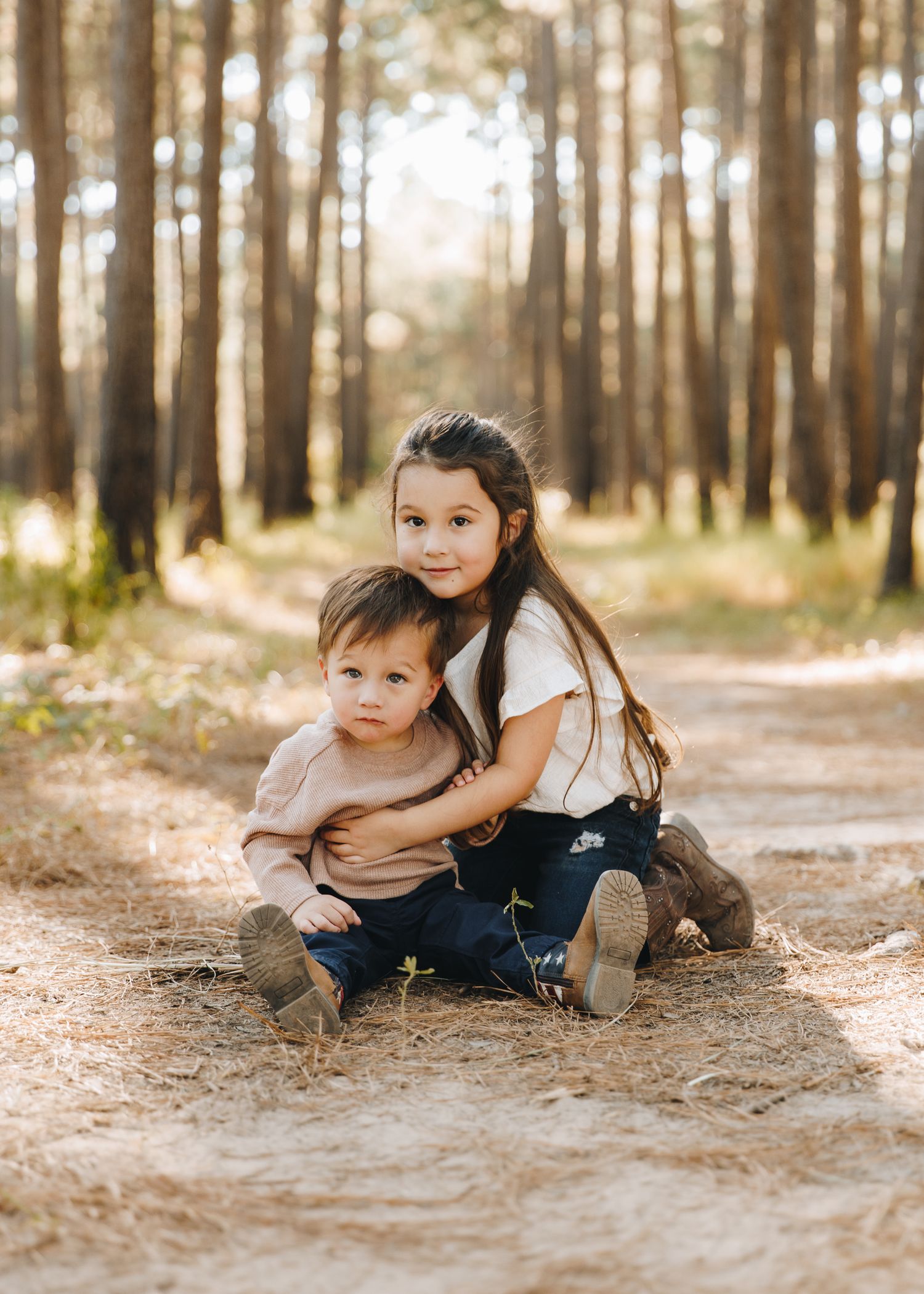Young girl hugging little brother while sitting on pine forest dirt path.