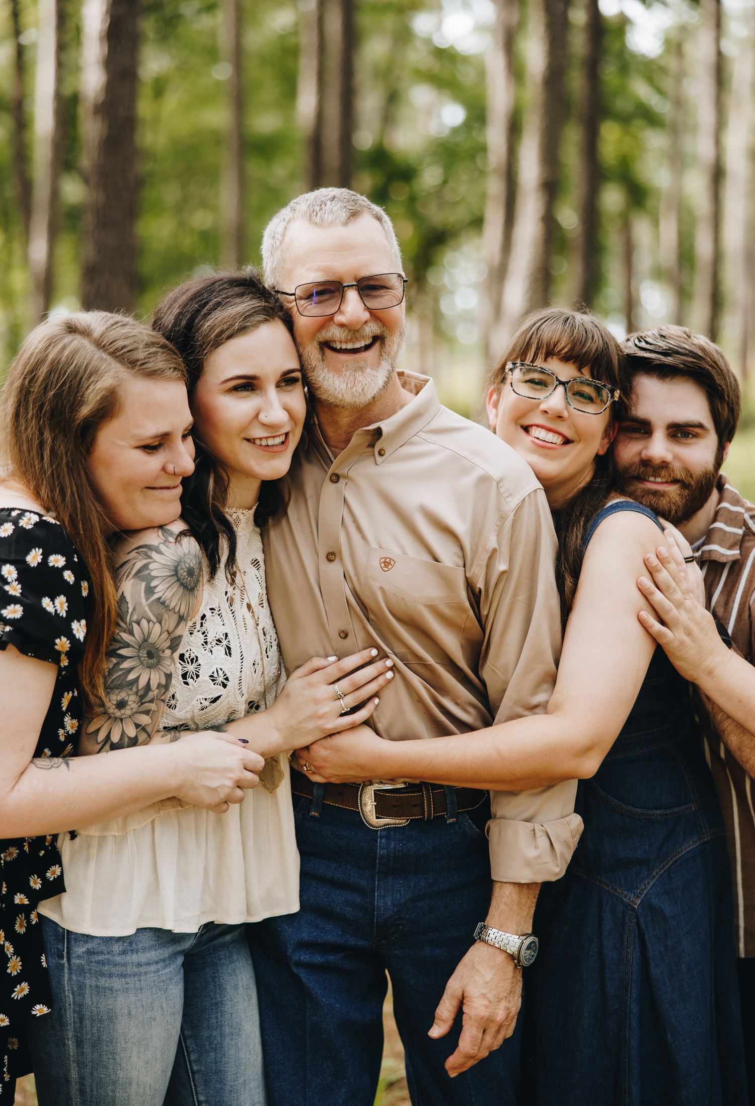 A happy multigenerational family hugging outdoors among tall pine trees during a warm family portrait session.