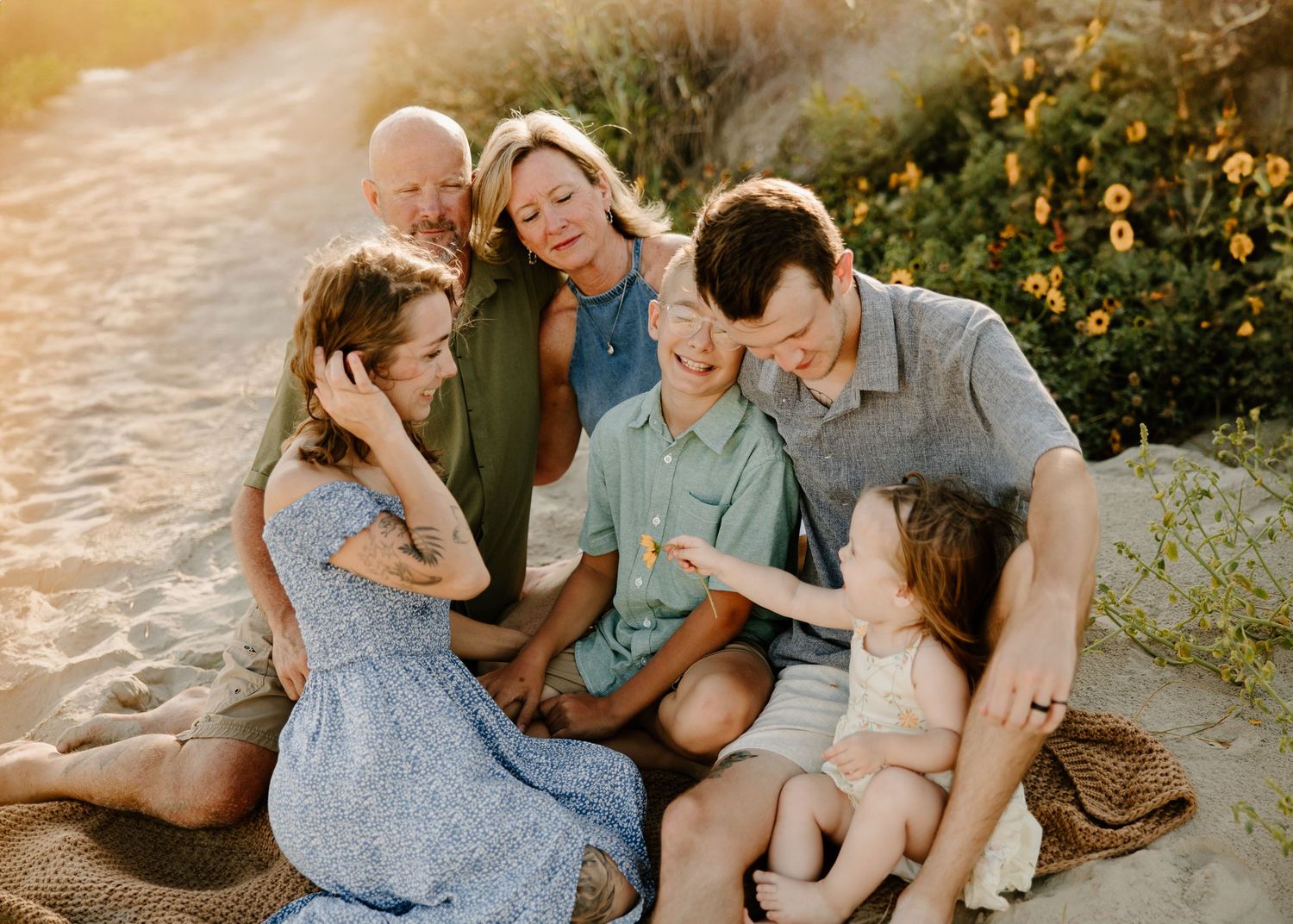 A loving family of six sits together on a sandy beach path surrounded by golden sunlight and flowers.