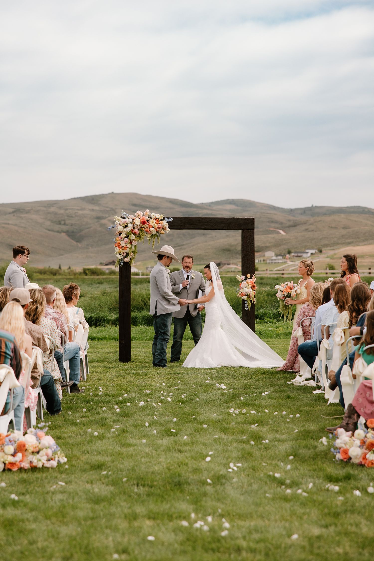 Outdoor wedding ceremony with couple at floral arch, rolling hills backdrop, and guests seated on both sides.