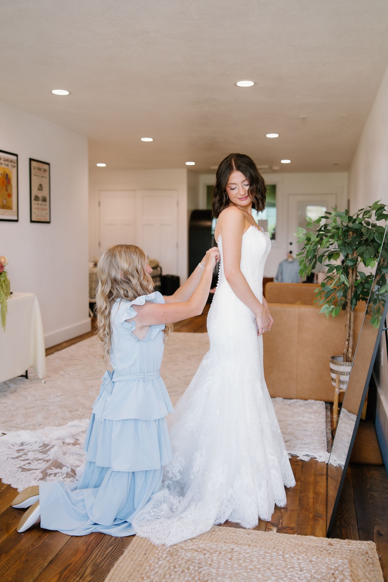 Young flower girl in blue dress helping bride button her fitted lace wedding gown.