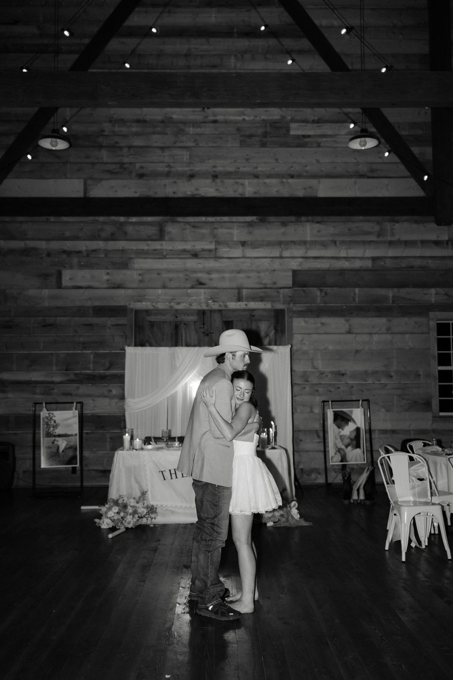 Bride and groom share first dance in rustic wooden barn with string lights overhead.