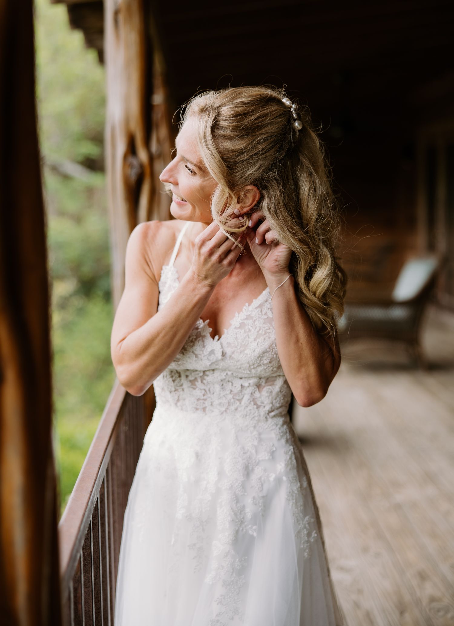 Bride in lace wedding gown adjusting earring on rustic wooden porch.