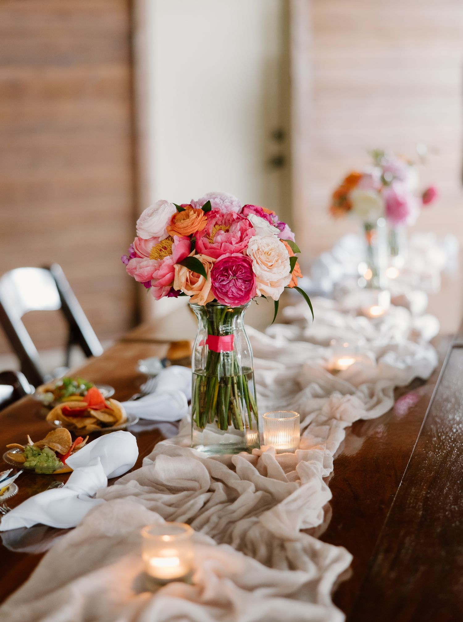Elegant wedding reception table with pink and orange floral centerpieces and candles on a linen runner.