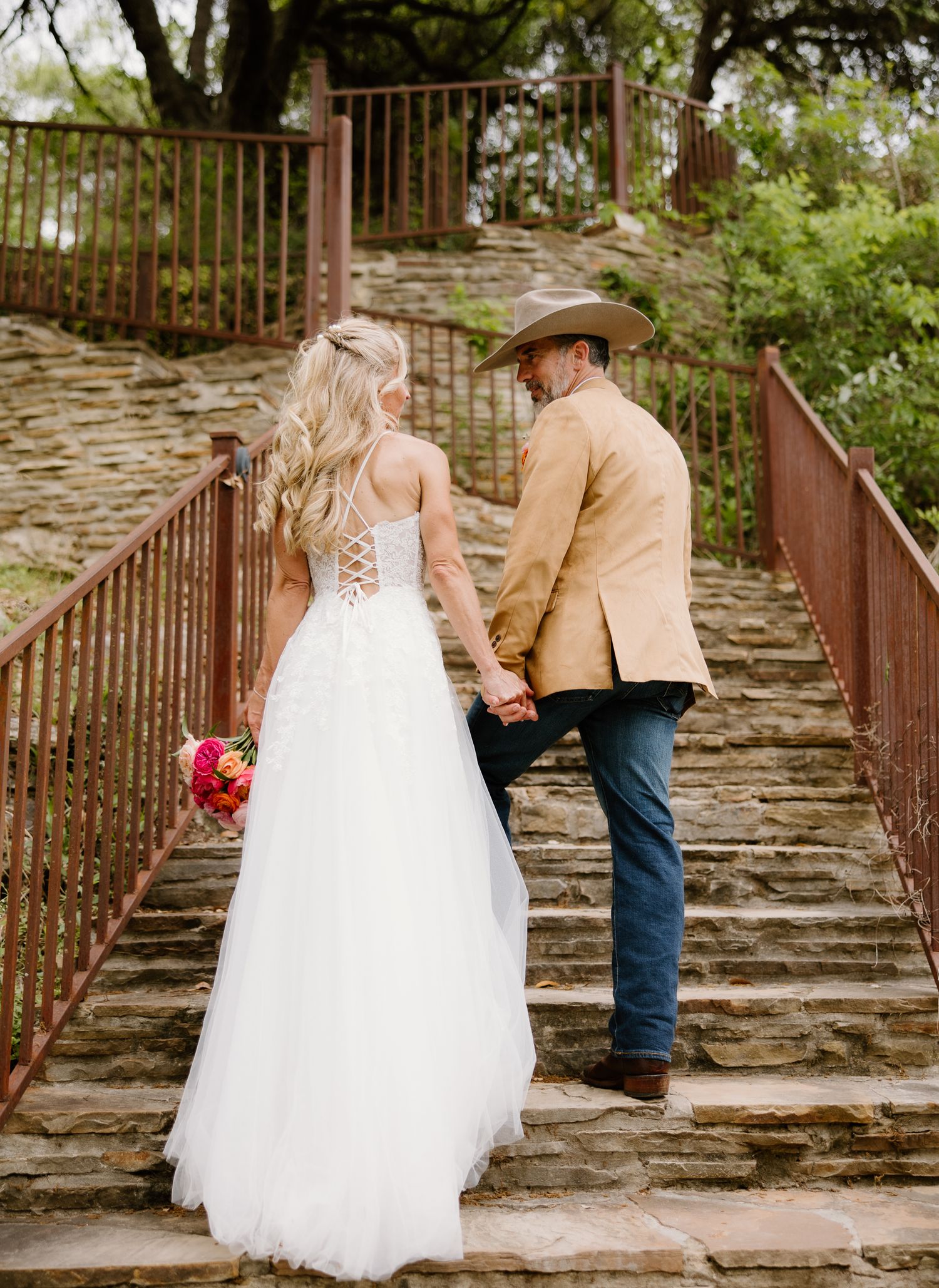 Bride and groom holding hands walking up stone steps, groom wearing cowboy hat and tan blazer.