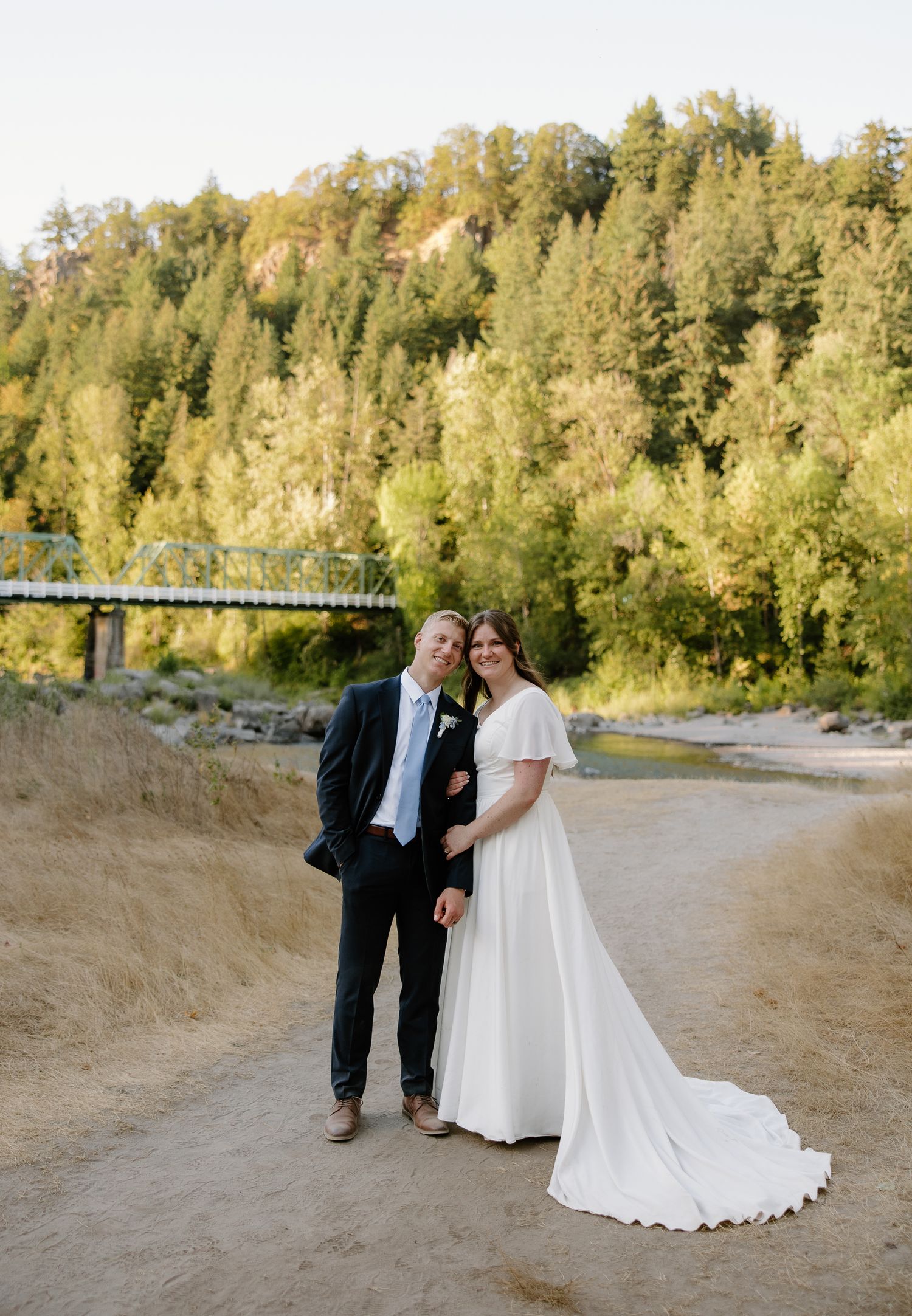 Smiling bride in white flutter-sleeve gown and groom pose on sandy path with river and forest behind.