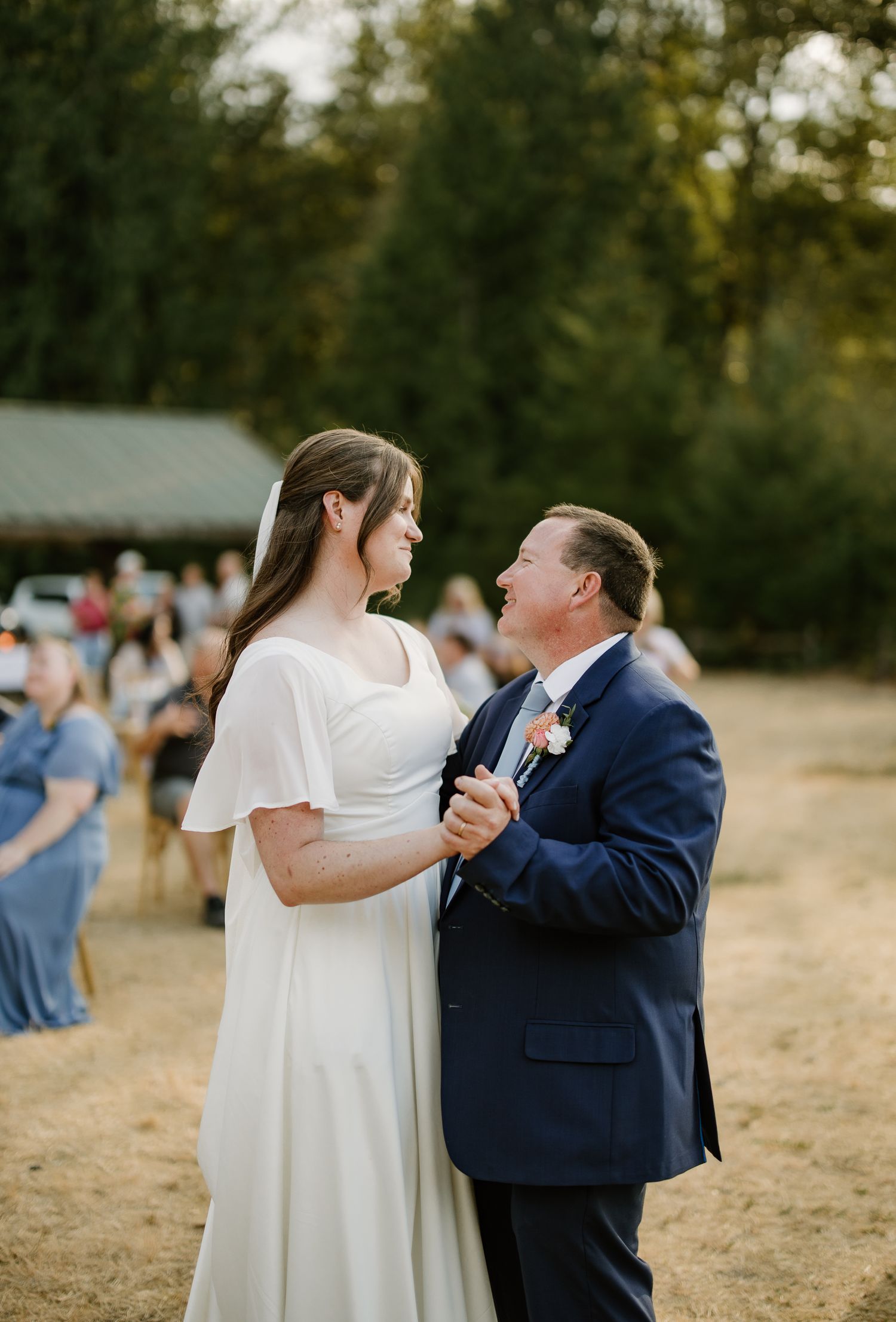 Bride dances with older man in navy suit at outdoor wedding reception at sunset.