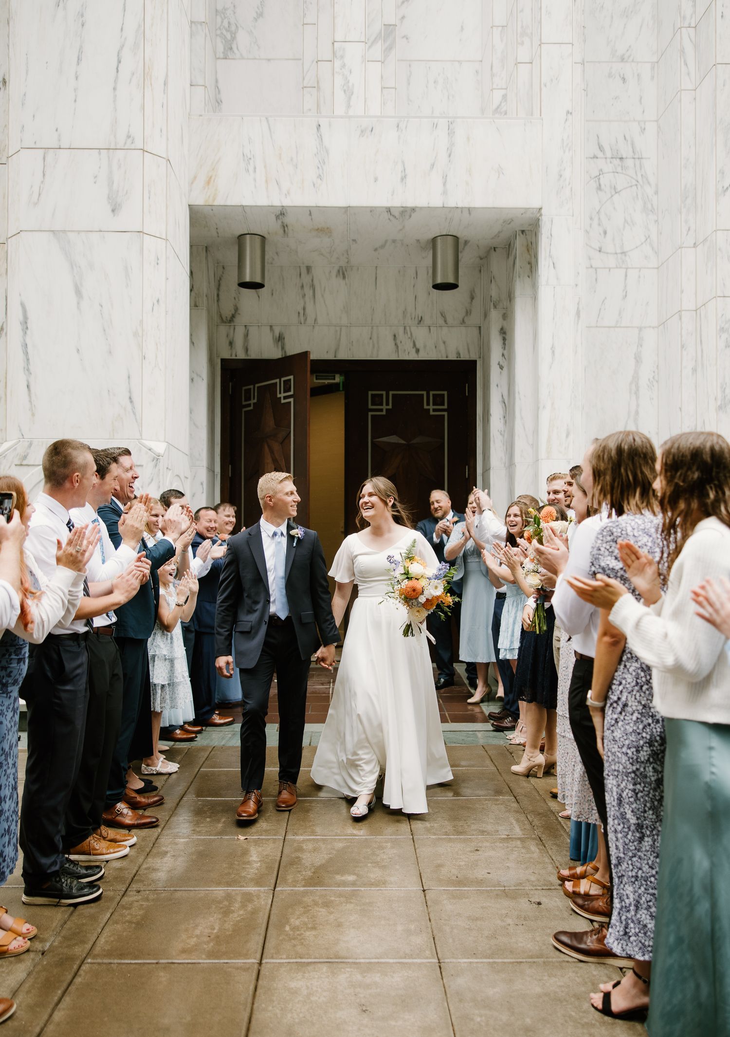 Bride and groom walk through cheering guests in a marble hall after their wedding ceremony.