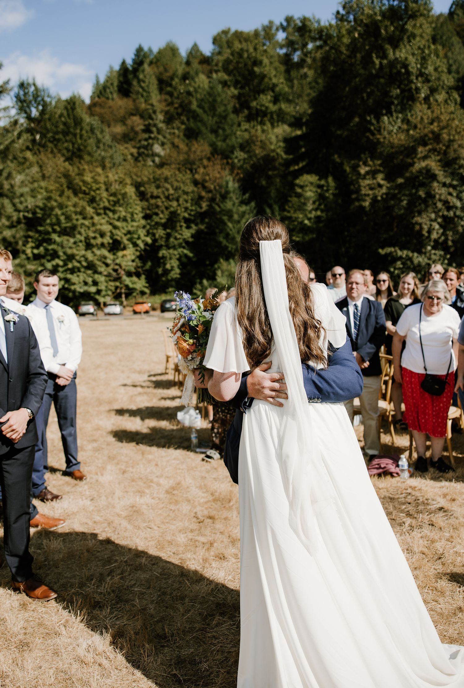 Bride walking down outdoor aisle in white gown with veil, escorted by family member holding colorful bouquet.