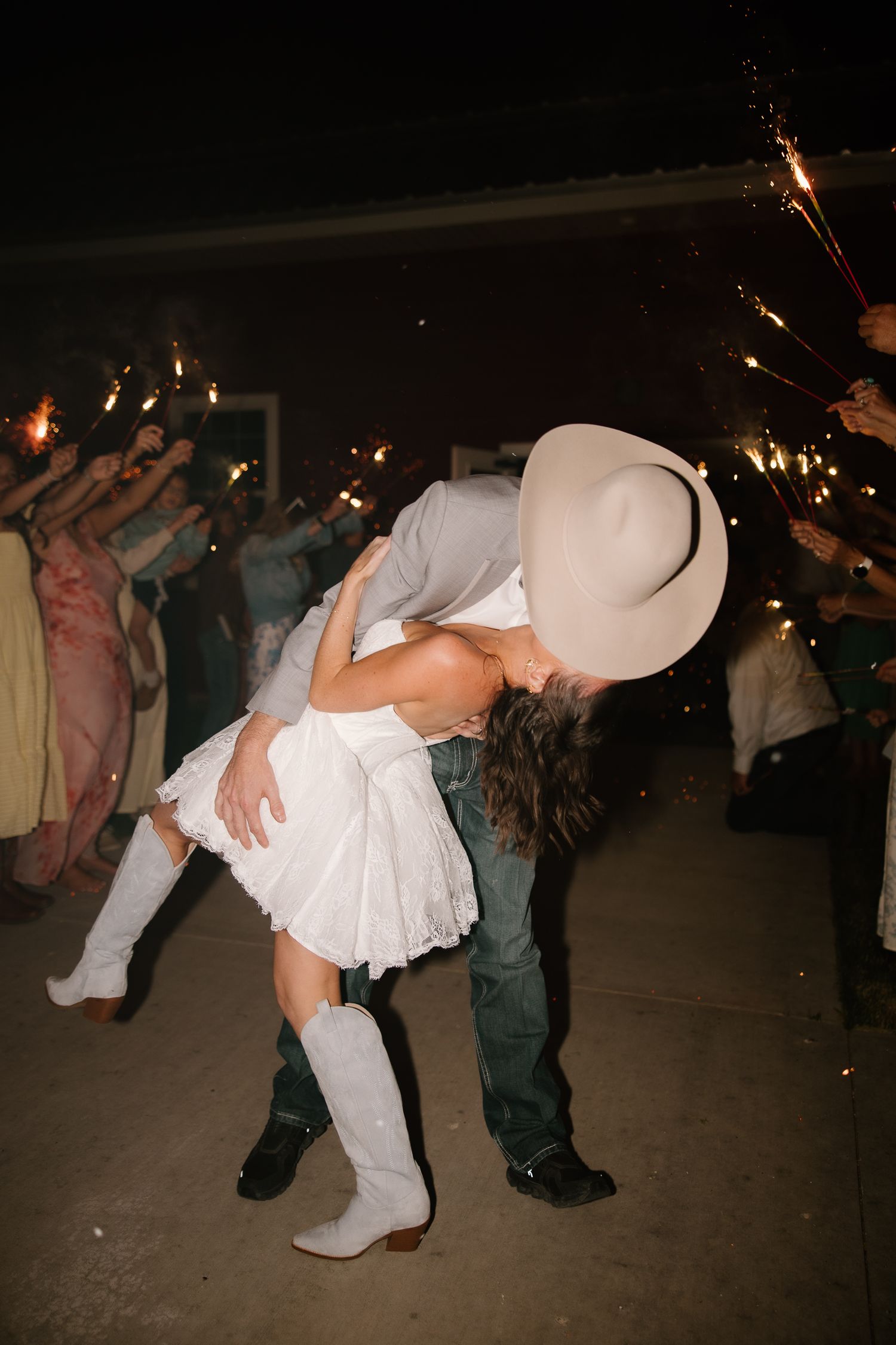 Bride and groom share romantic dip kiss during sparkler send-off exit at outdoor country wedding celebration at night.