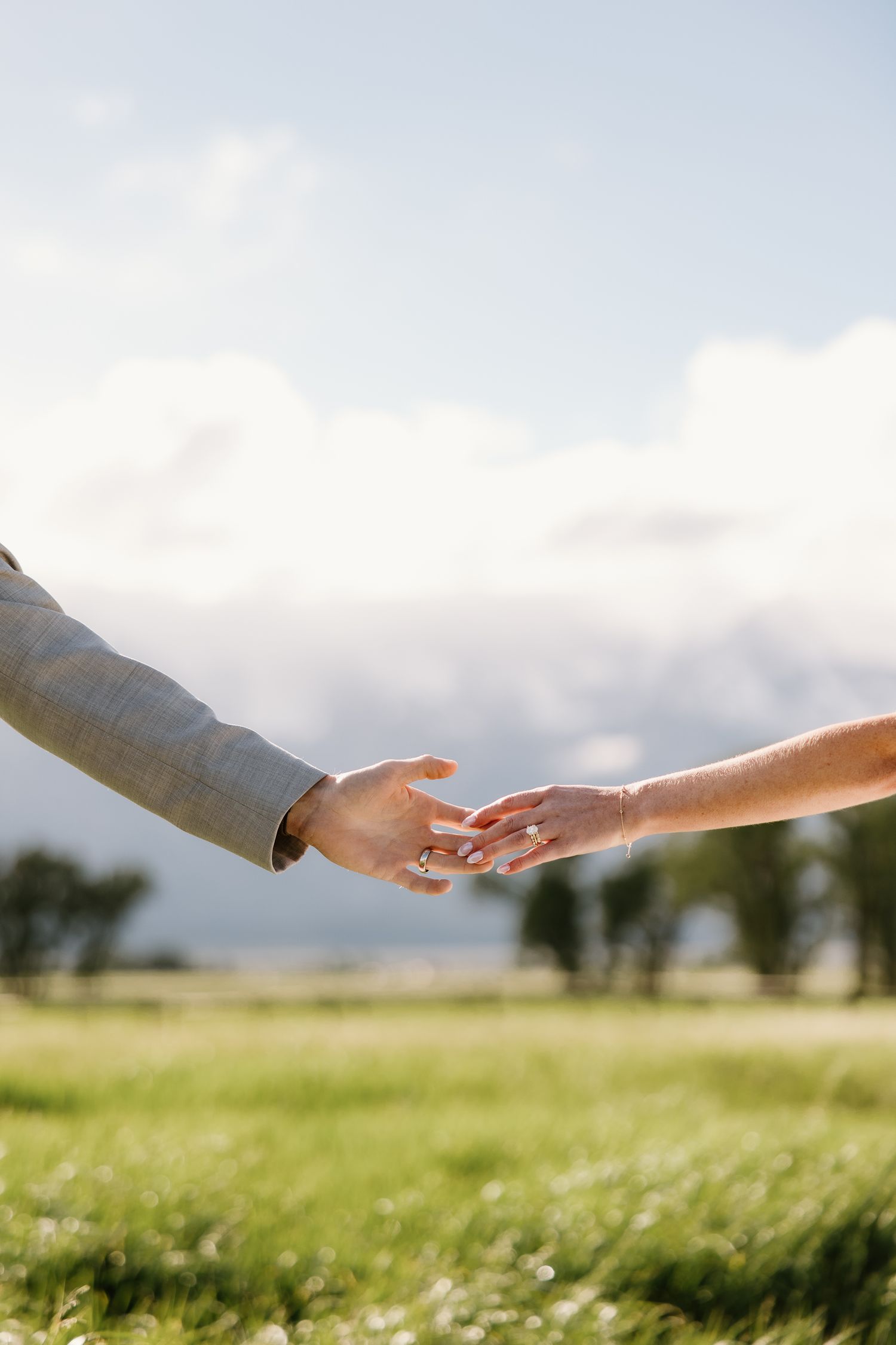 Close-up of bride and groom gently touching hands in a sunlit green meadow with wedding rings visible.