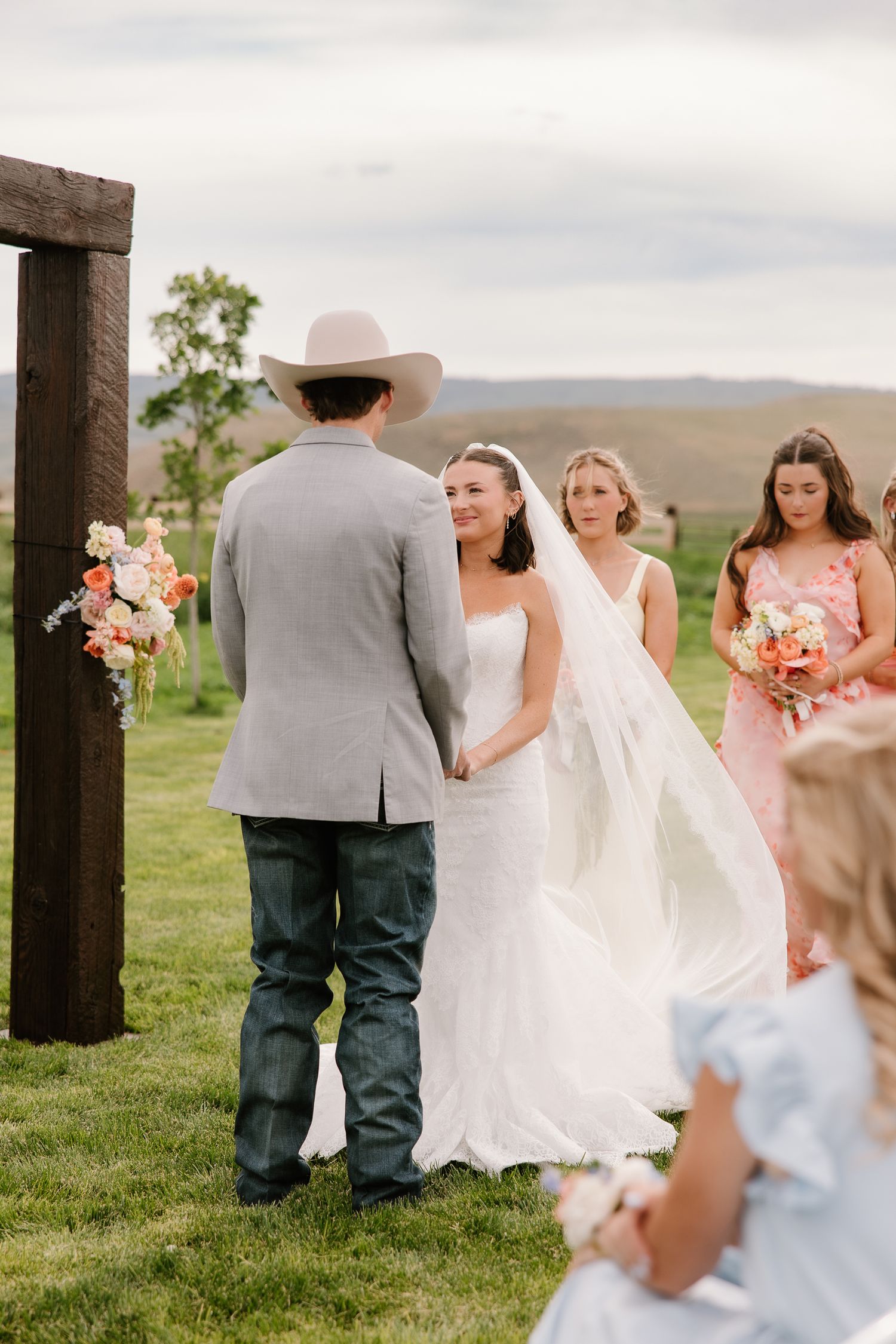 Smiling bride in white gown holds grooms hands at outdoor ceremony altar with bridesmaids nearby.