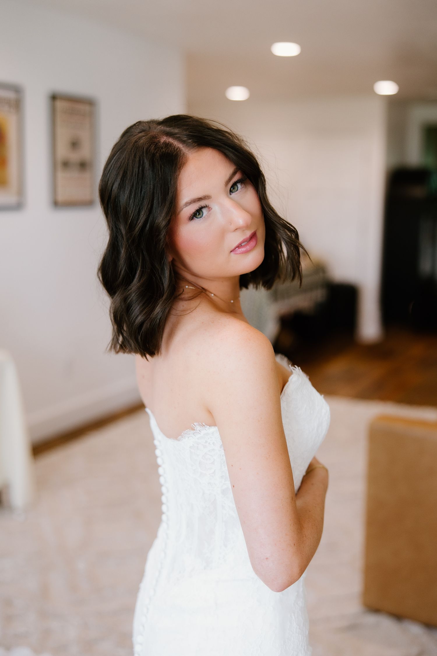 Bride in strapless white lace wedding dress with short dark wavy hair looking over shoulder indoors.