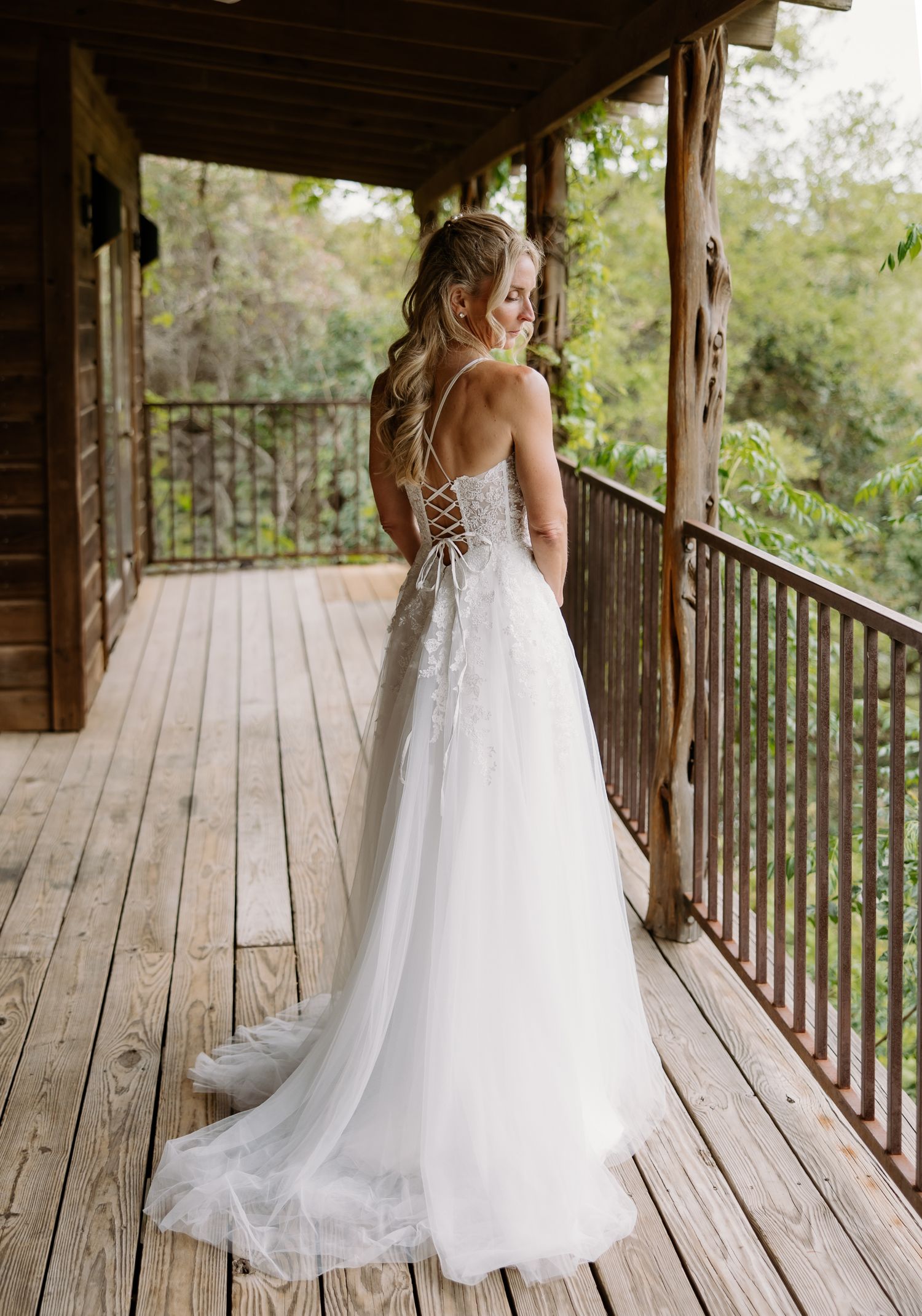 Bride in elegant lace-up back wedding gown with tulle skirt on rustic wooden porch surrounded by lush greenery.