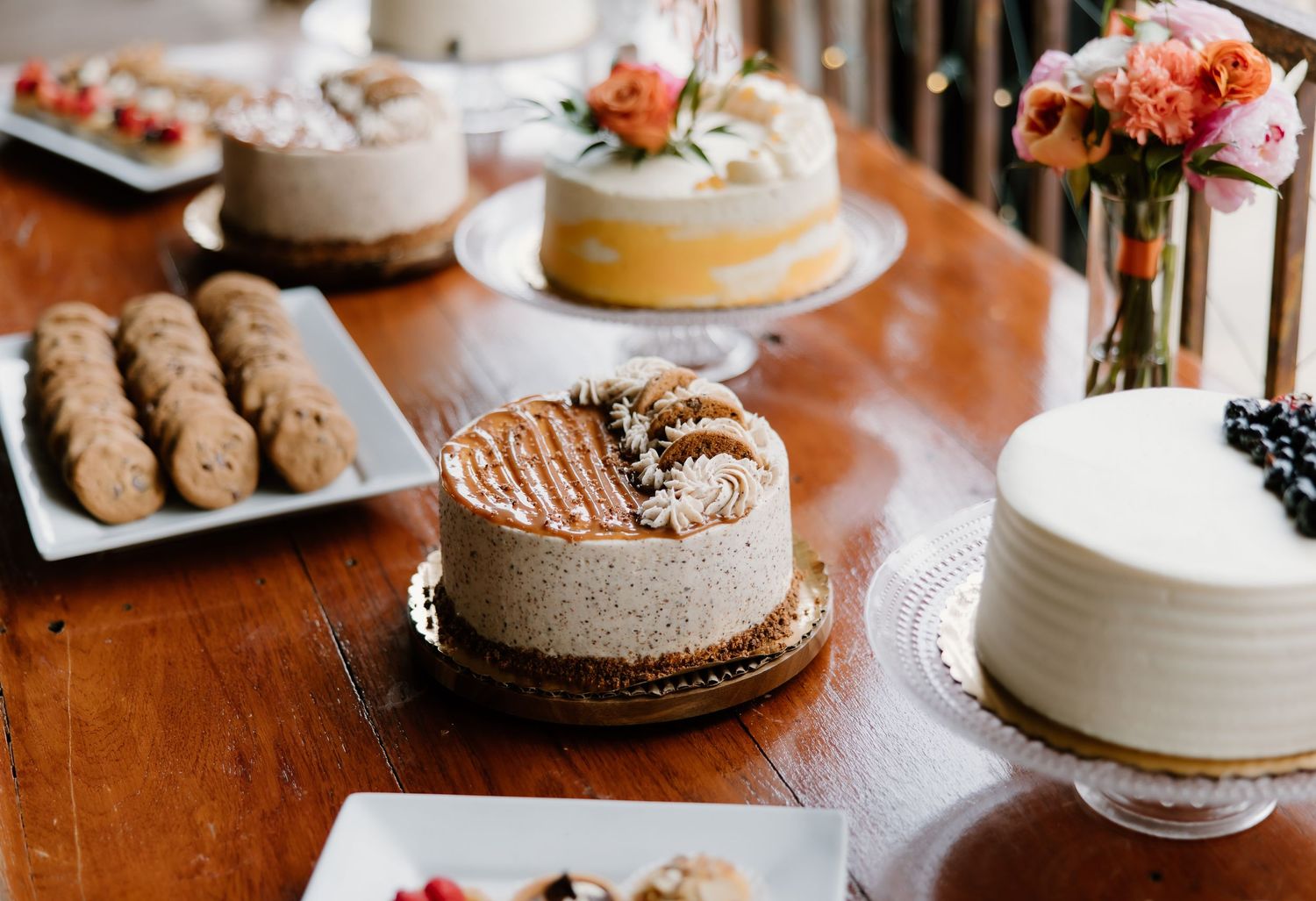 Elegant dessert table with assorted cakes, cookies, and floral arrangements on a wooden surface.