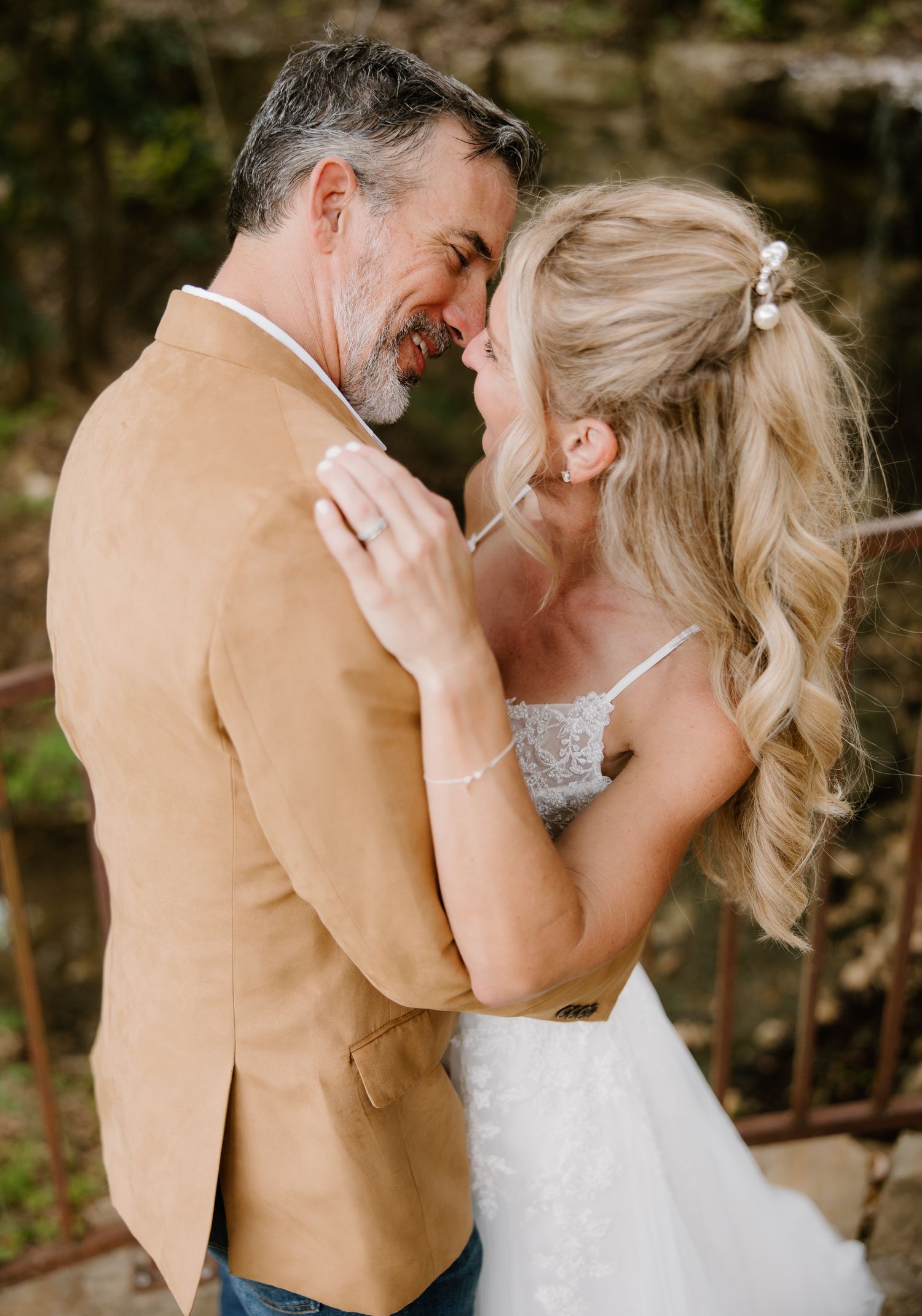 Newlywed couple in romantic embrace, bride in lace gown and groom in tan jacket, touching foreheads.