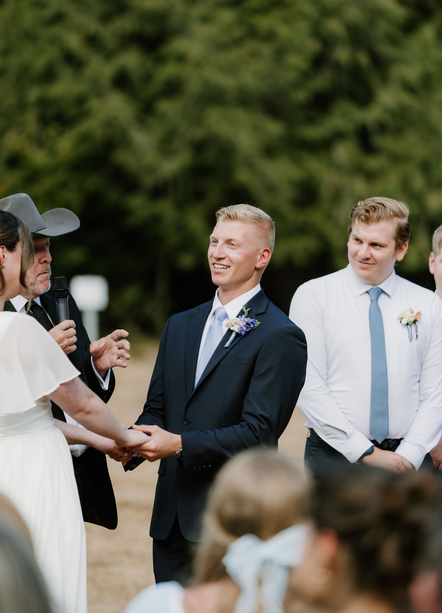 Smiling groom in navy suit holds hands with bride during outdoor wedding ceremony with groomsmen.