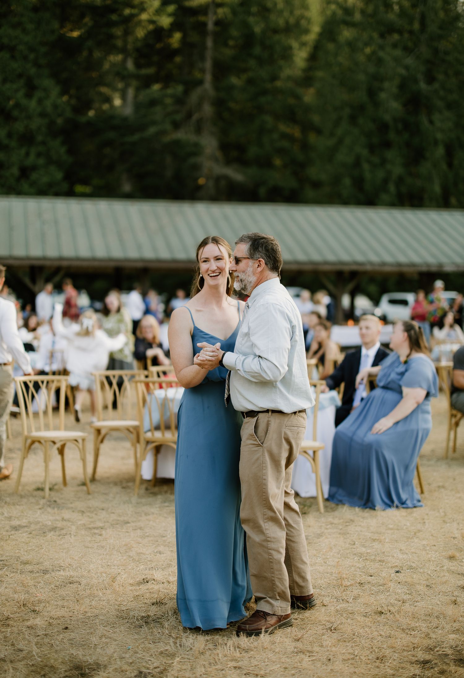 Bridesmaid in blue gown dances with older man at outdoor wedding reception on sandy ground.