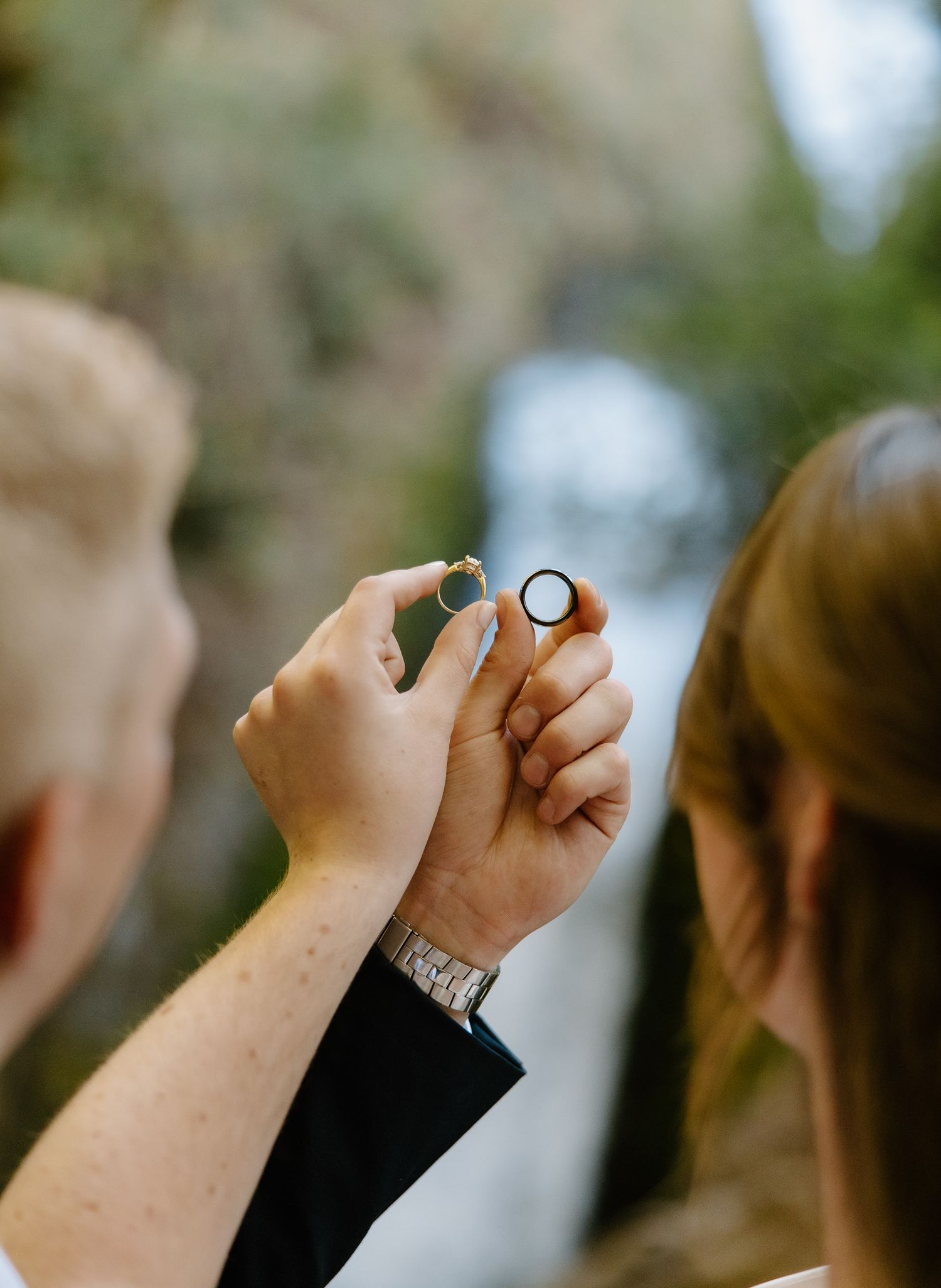 Couple exchanging wedding rings outdoors with a scenic waterfall in the soft-focus background.