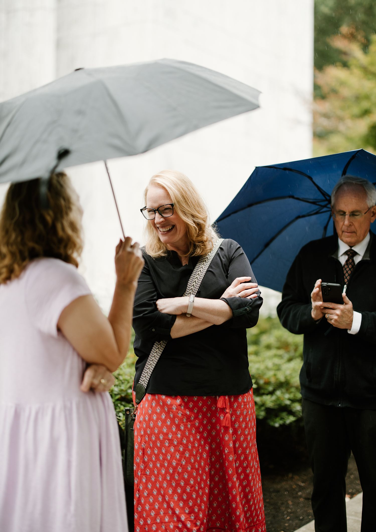 Guests laugh and chat under umbrellas on a rainy wedding day outside a white building.