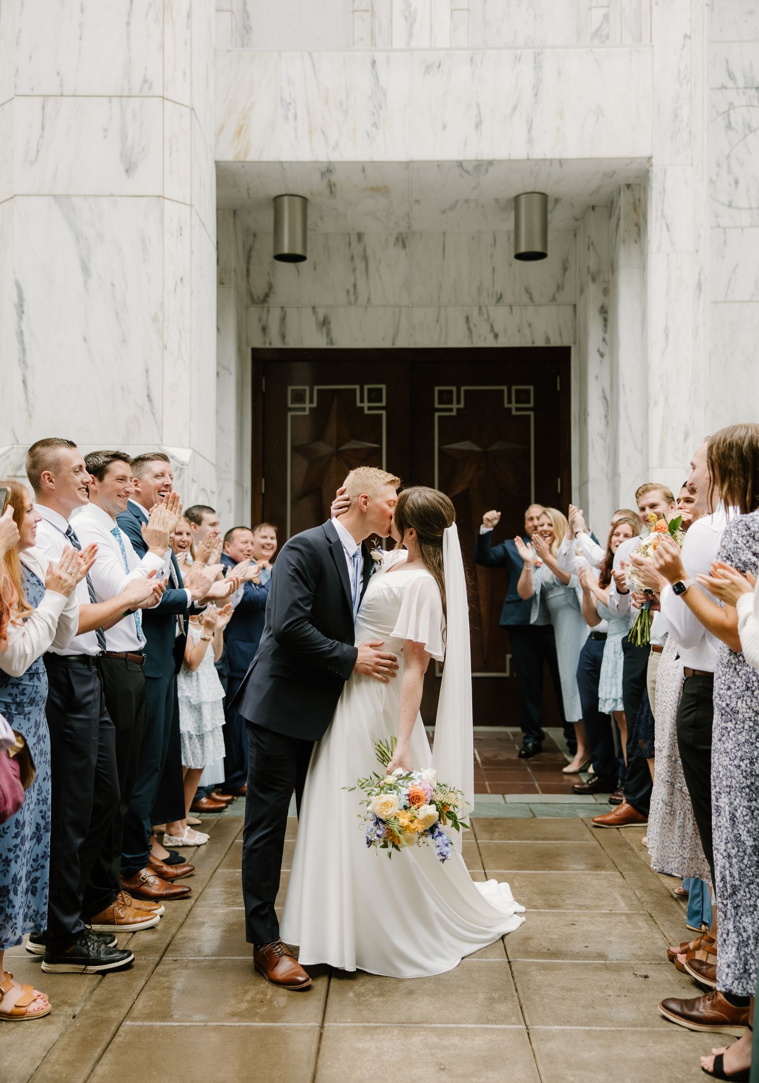 Bride and groom share a kiss while wedding guests clap and cheer outside a marble building entrance.