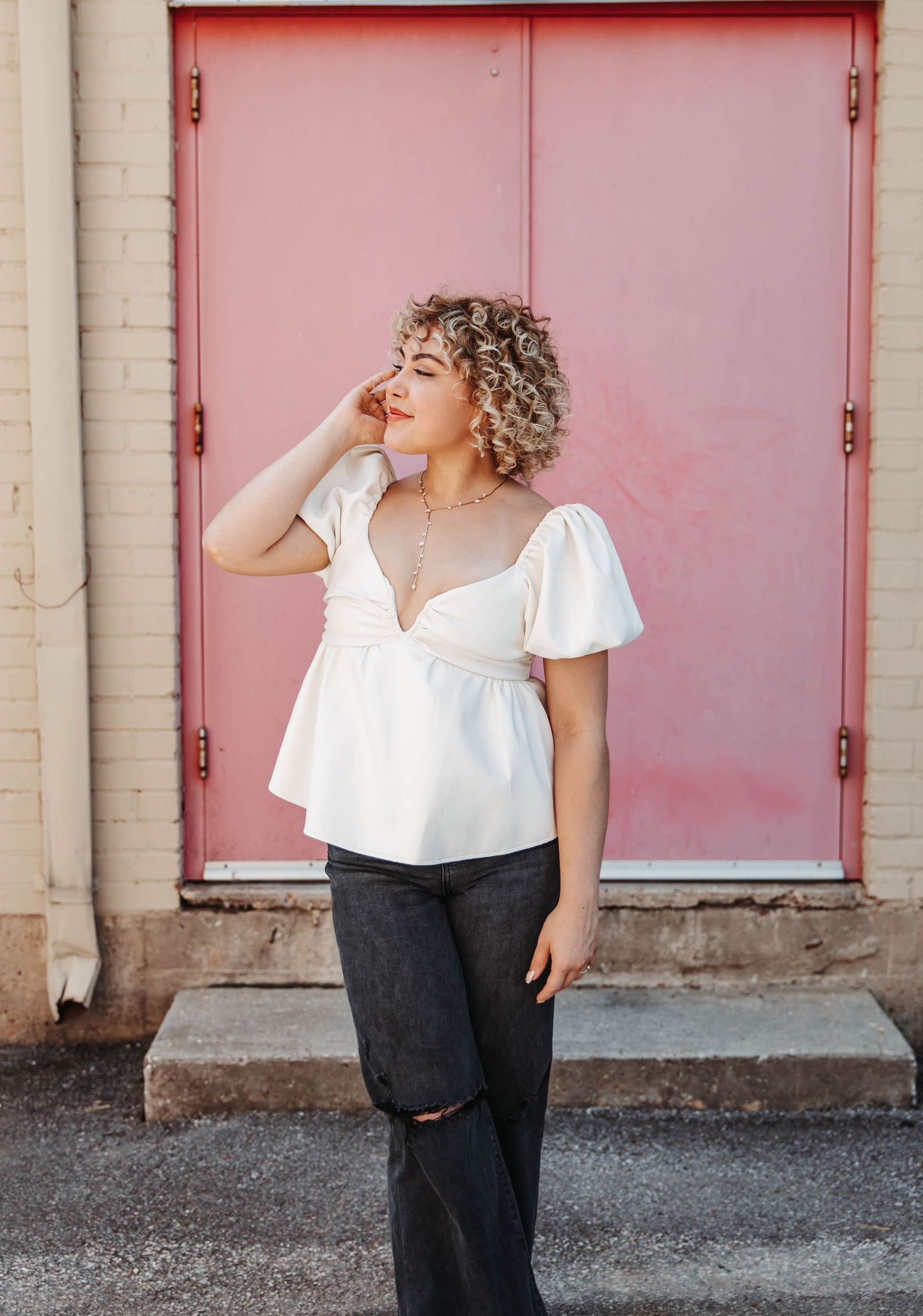 Young woman in white puff-sleeve top and dark jeans poses in front of a pink door.