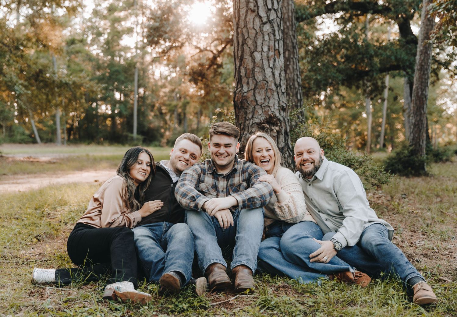 Group of five adults laughing and sitting together at the base of a large tree in a wooded area.