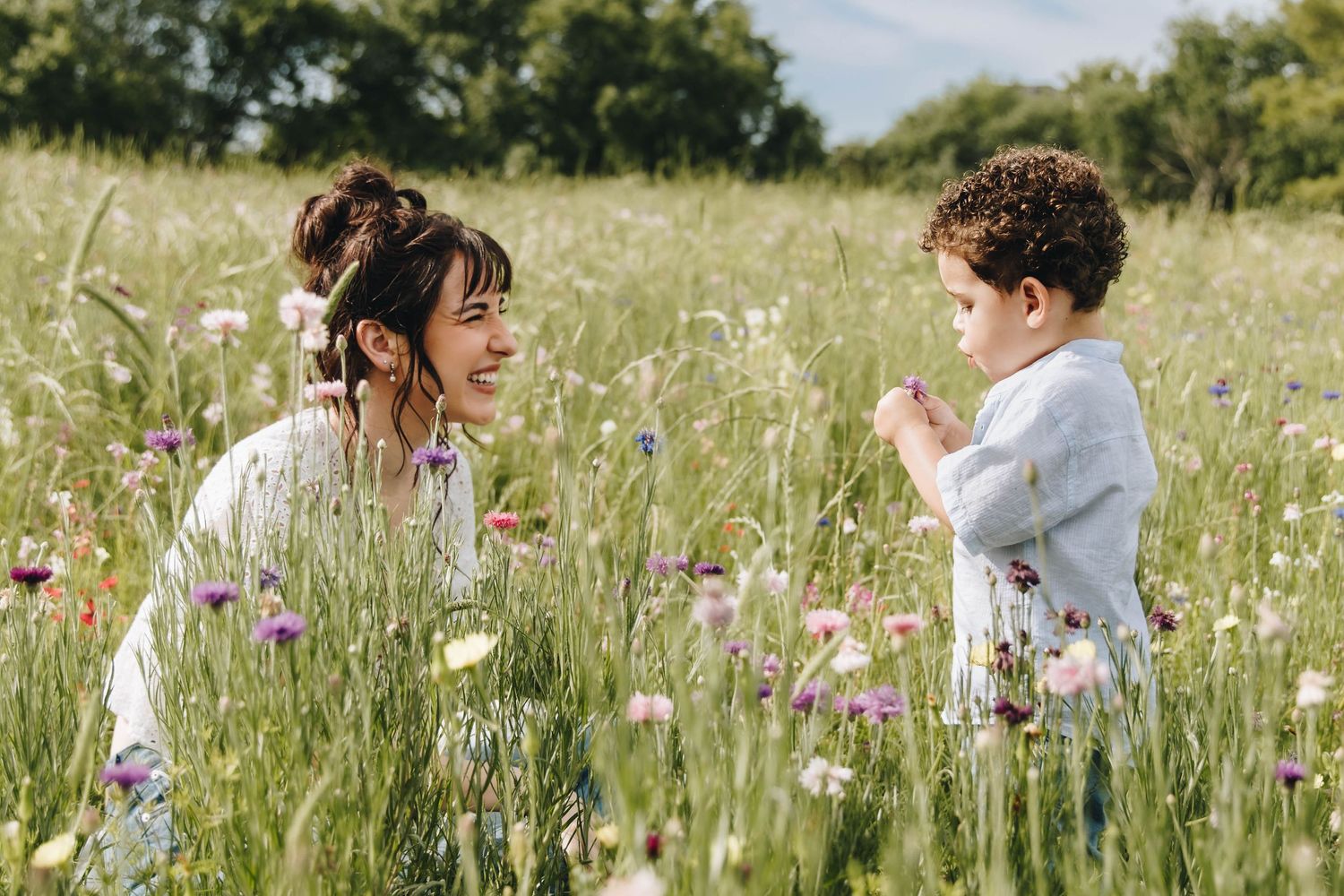 Smiling mother kneels in wildflower meadow facing young curly-haired toddler on sunny day.