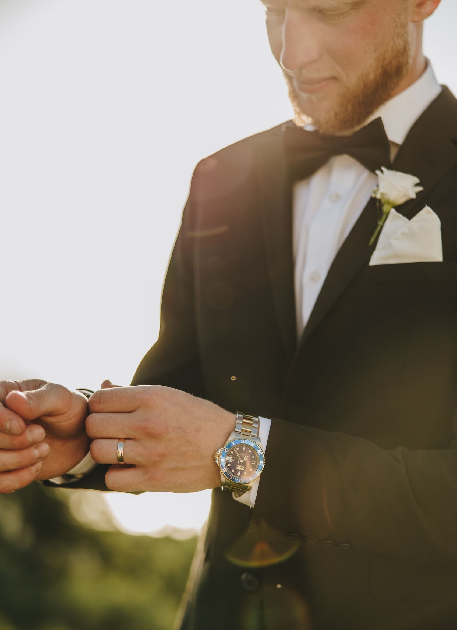 Groom in black tuxedo with bow tie and boutonniere adjusting cufflinks, wearing a luxury watch and wedding ring.