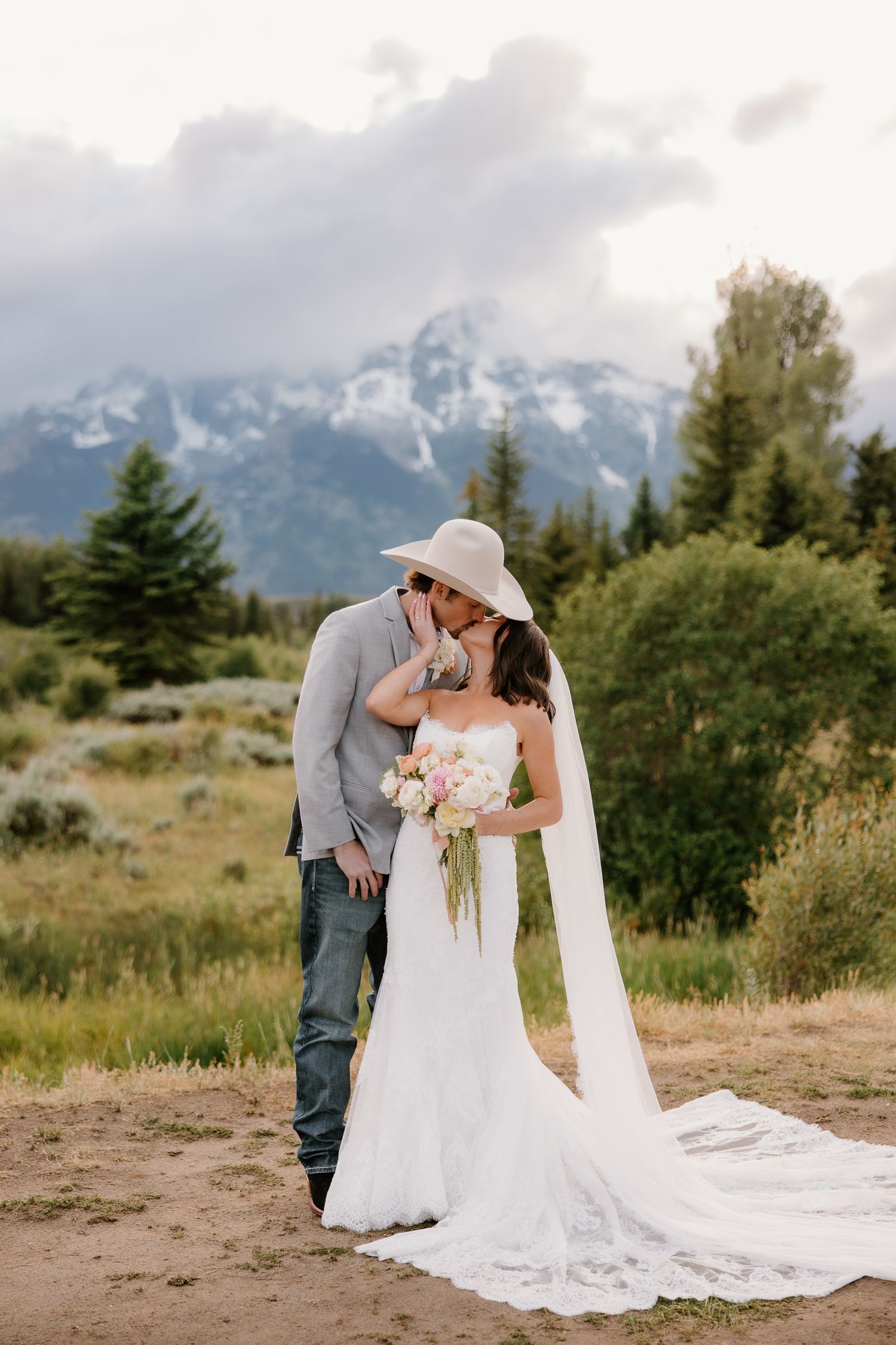 Bride and groom share a kiss with stunning Grand Teton mountains backdrop, bride holding pink and white bouquet.