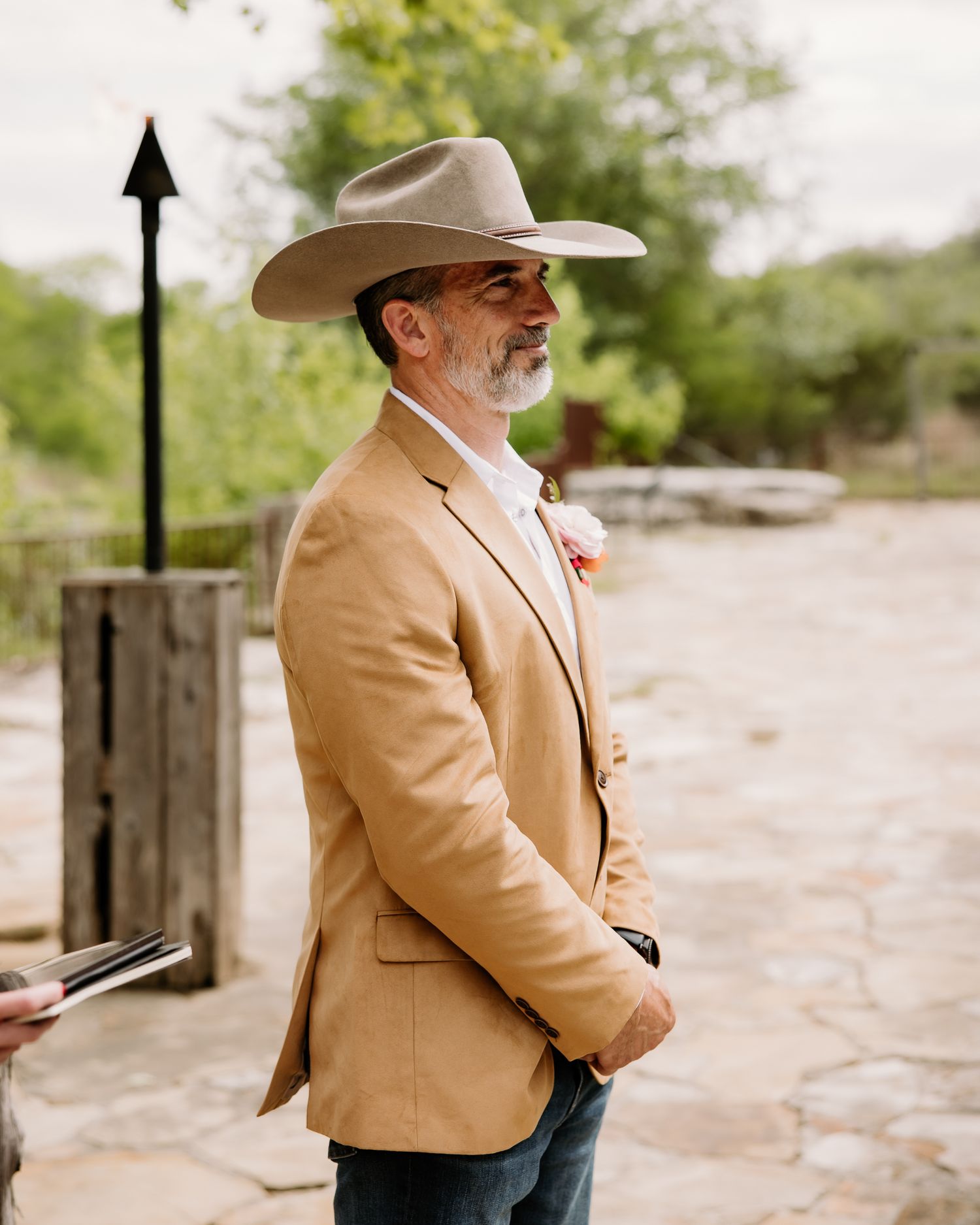 Man in a tan blazer and cowboy hat wearing a boutonniere at an outdoor wedding ceremony.