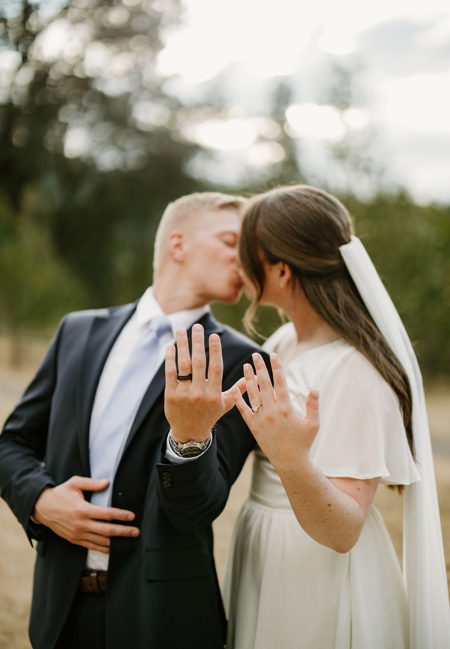Newlyweds kissing while showing off wedding rings, bride in white gown with veil, groom in dark suit with blue tie.