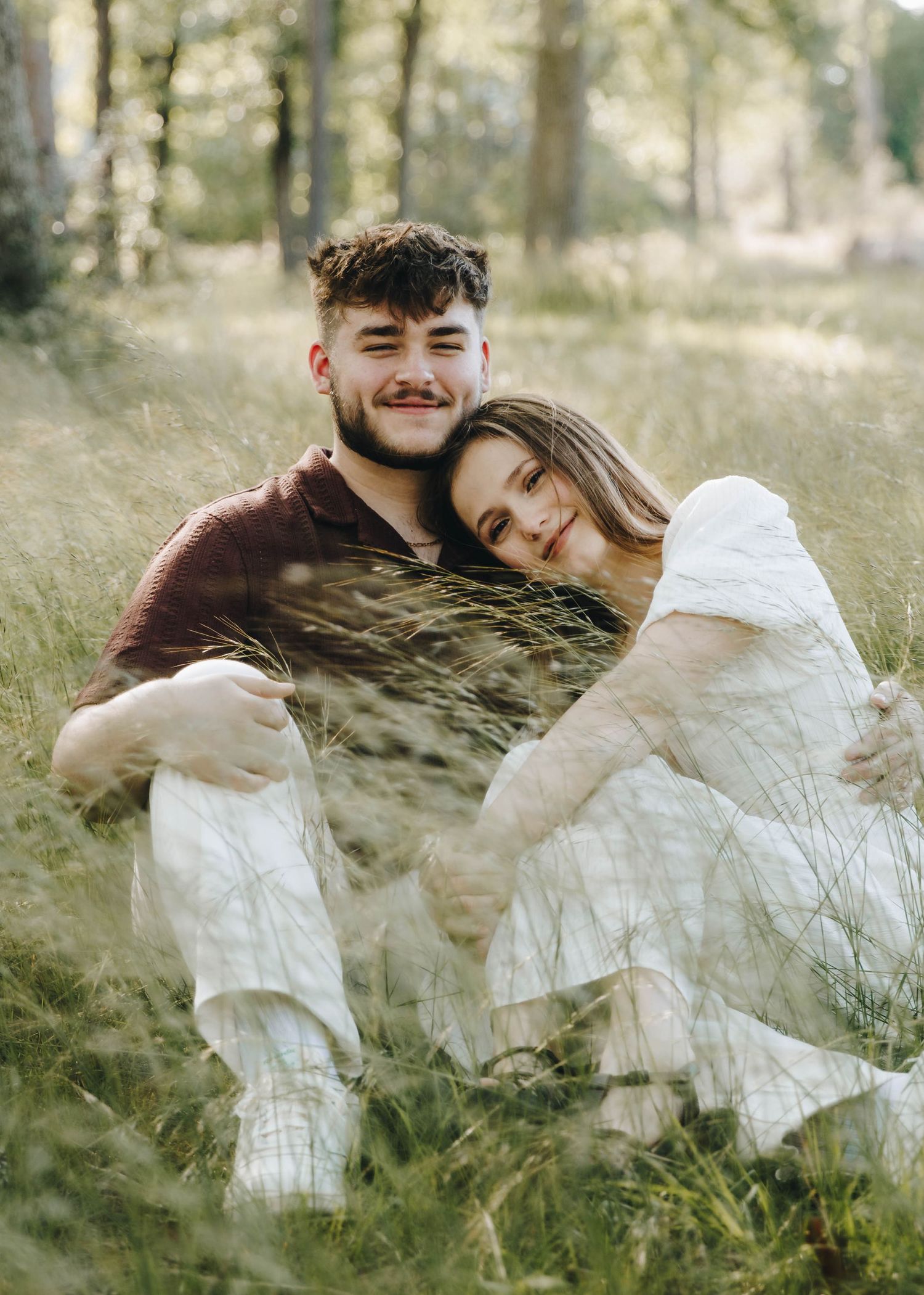 Couple sitting together in a grassy woodland meadow, woman leaning on mans shoulder.