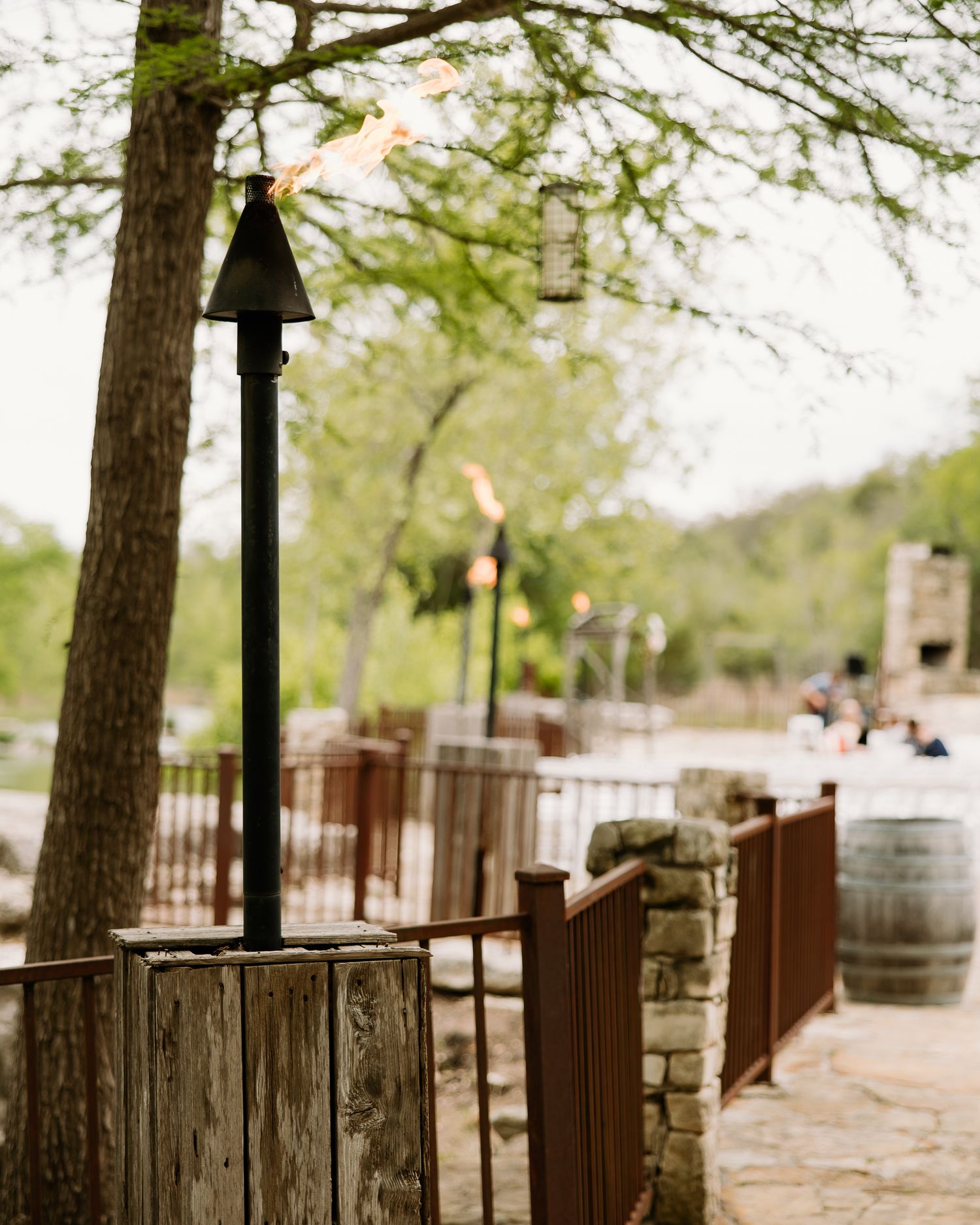 Lit tiki torch on stone post along rustic wooden fence at outdoor wedding venue with trees in background.