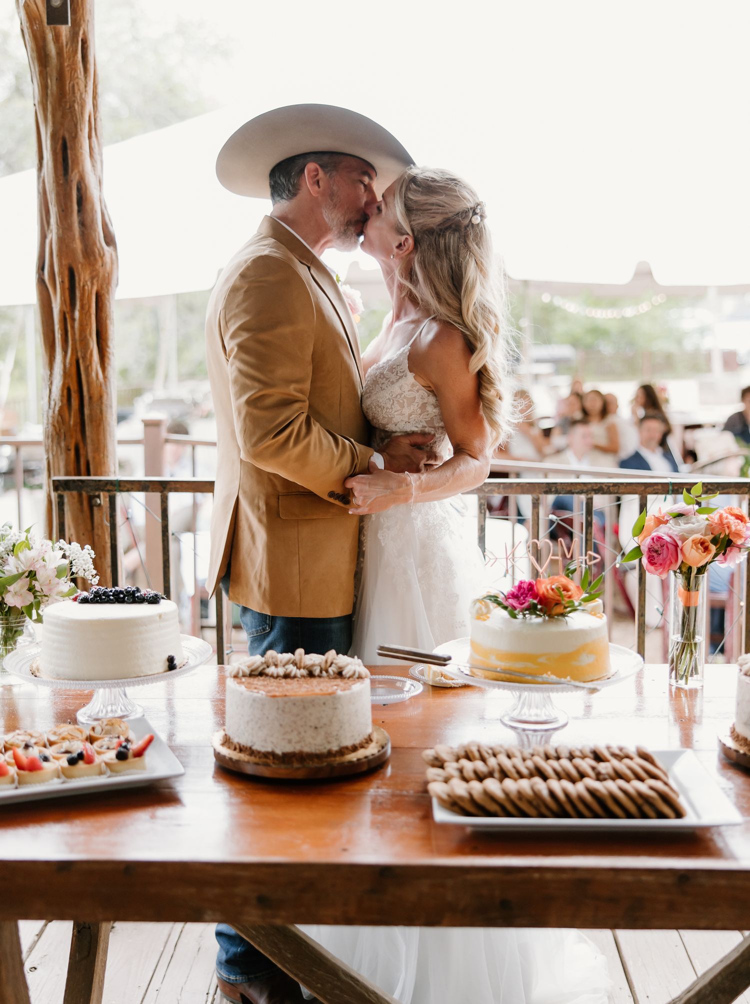Couple sharing a kiss at wedding dessert table, groom wearing cowboy hat and tan blazer.