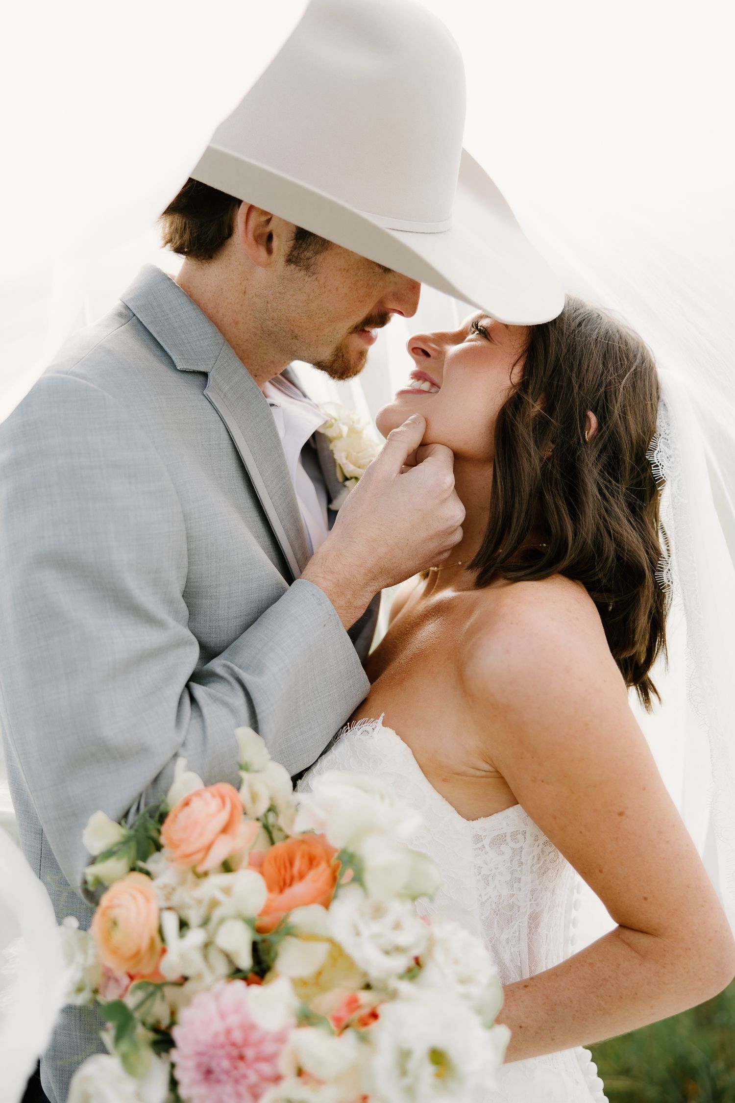 Groom in gray suit and cowboy hat tenderly holds brides chin before a kiss.