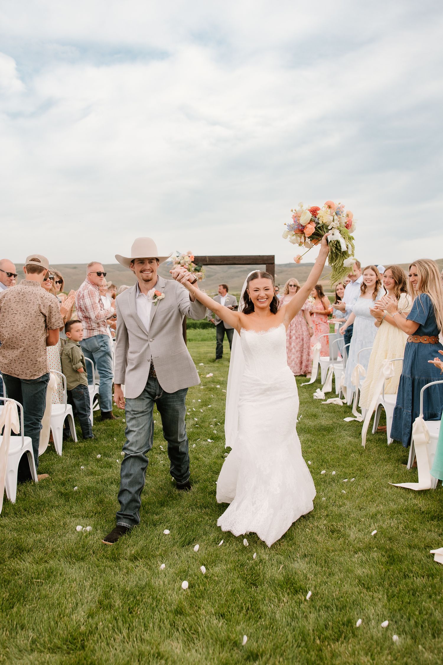Joyful bride and groom raise hands while walking back down the aisle after outdoor wedding ceremony.