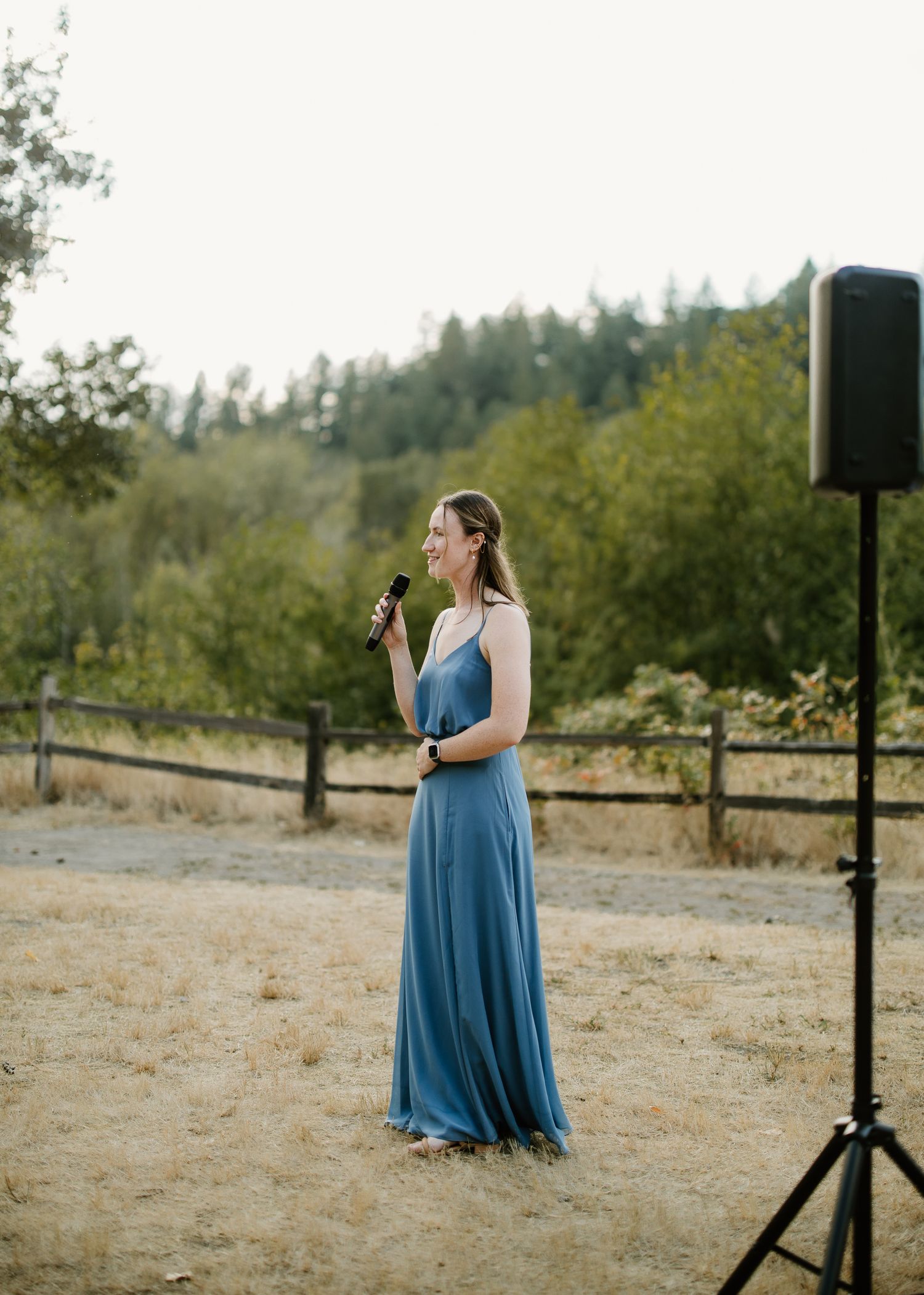 Bridesmaid in blue gown delivers speech at outdoor wedding with microphone and speaker.
