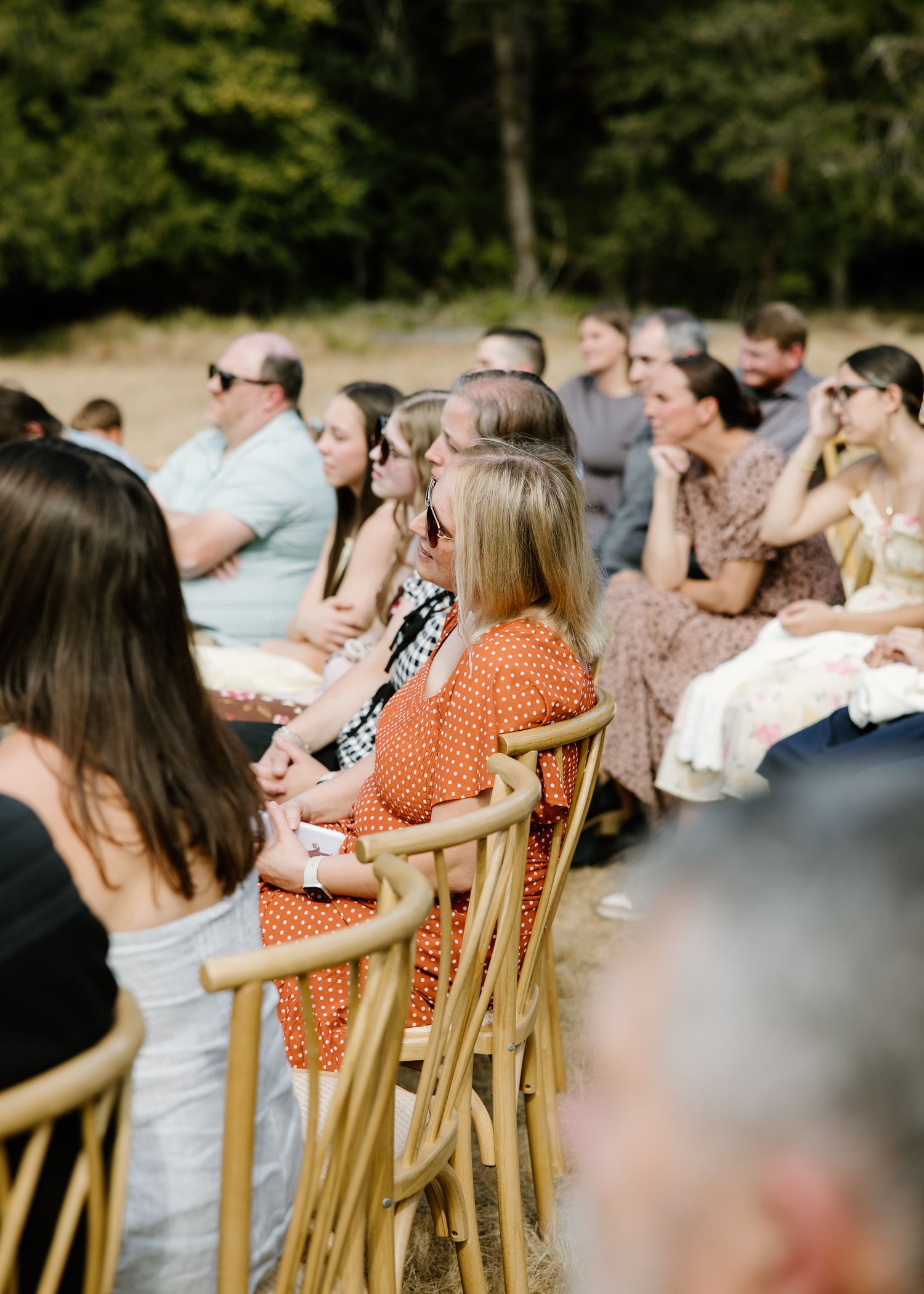 Wedding guests seated in wooden chairs watch outdoor ceremony surrounded by lush green forest.