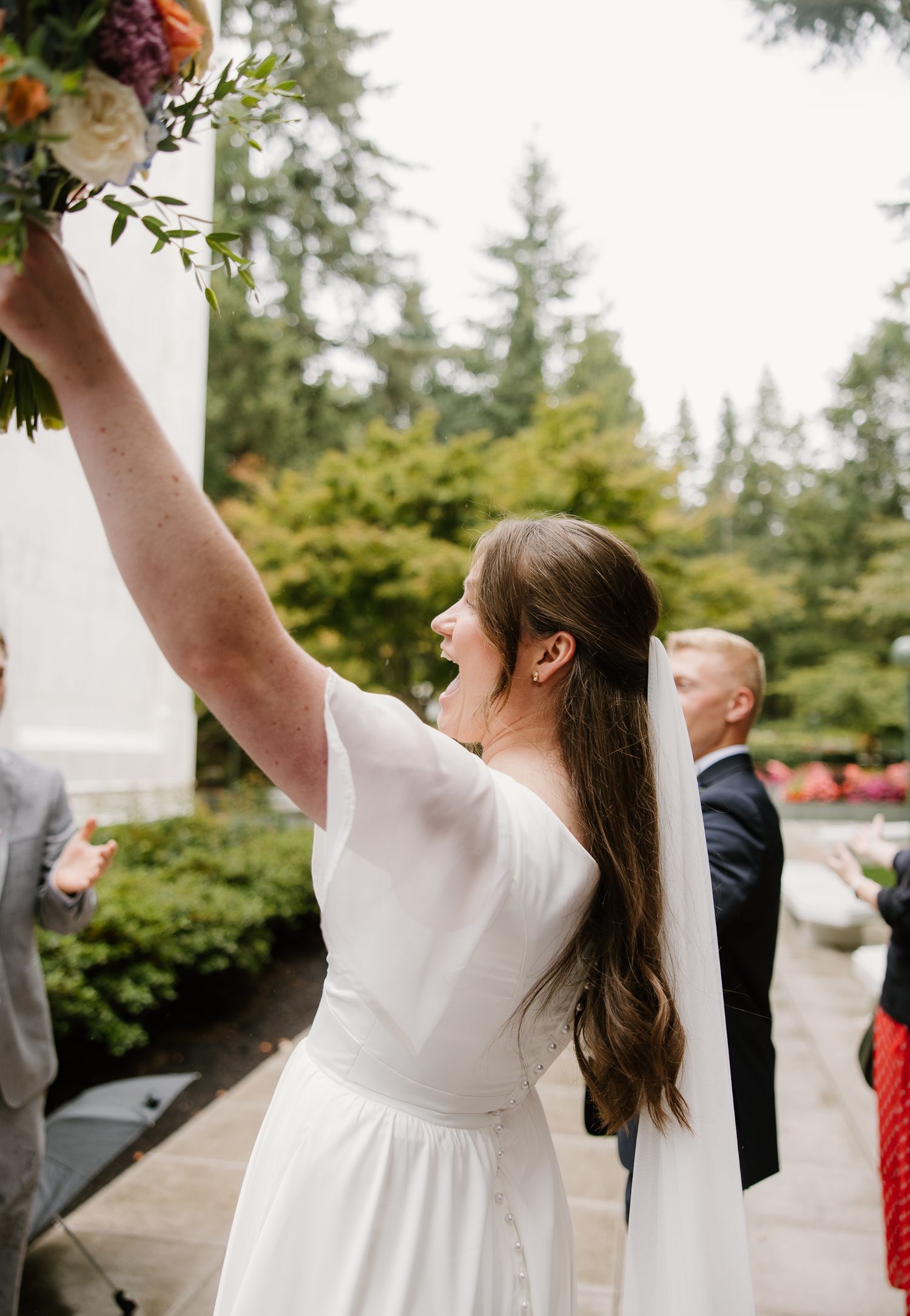 Joyful bride in white dress raising bouquet at outdoor wedding ceremony surrounded by lush greenery.