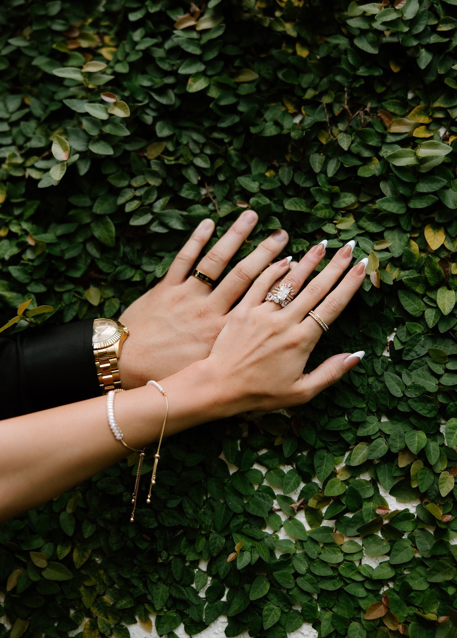 Close-up of bride and groom hands showing wedding rings, gold watch, and pearl bracelet against green ivy.