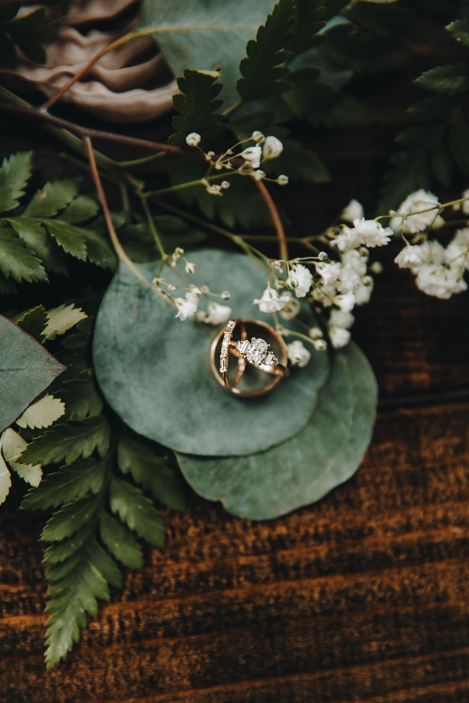 Gold wedding bands and diamond engagement ring nestled among green eucalyptus leaves and white baby's breath flowers.