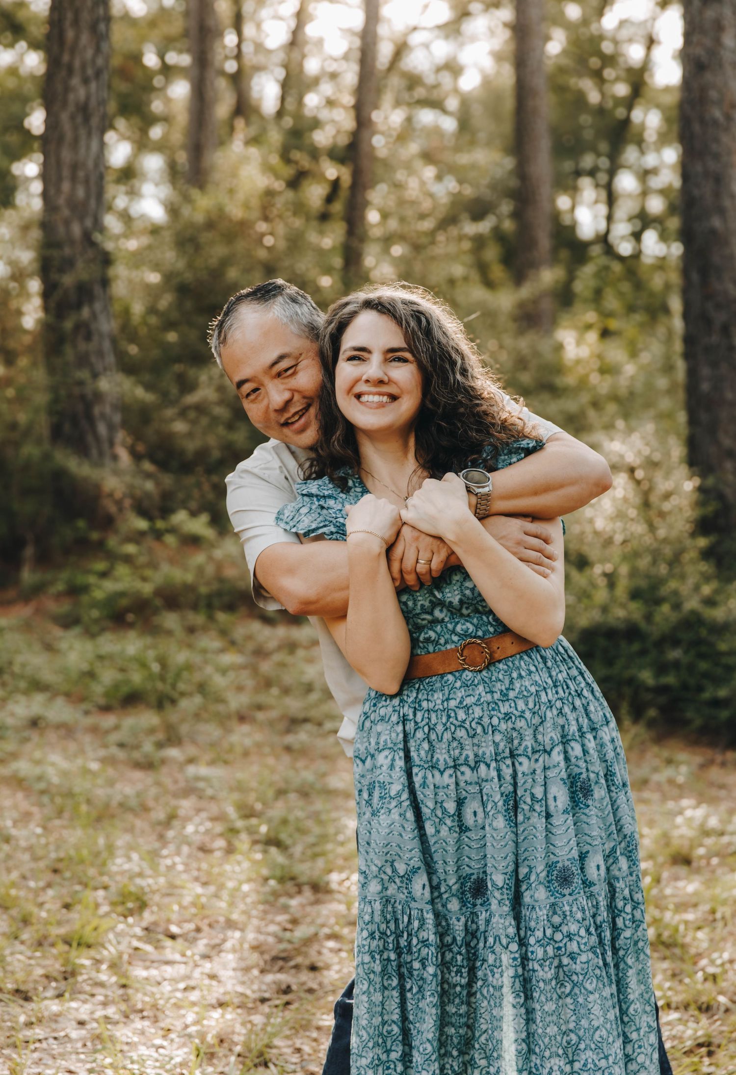 Man hugging woman from behind, both smiling in a sunlit forest setting.
