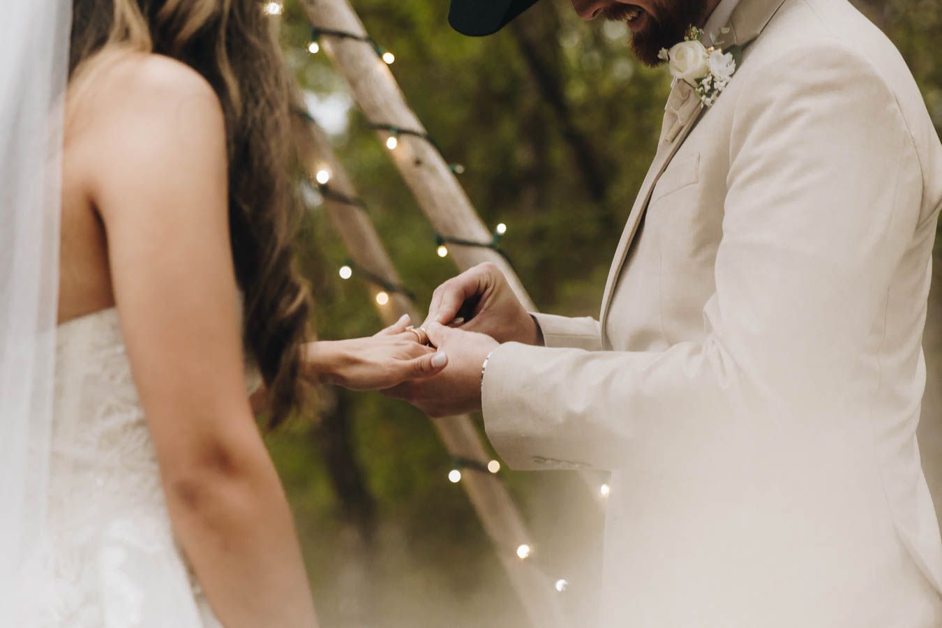 Groom places wedding ring on bride's finger during outdoor ceremony with string lights in the background.